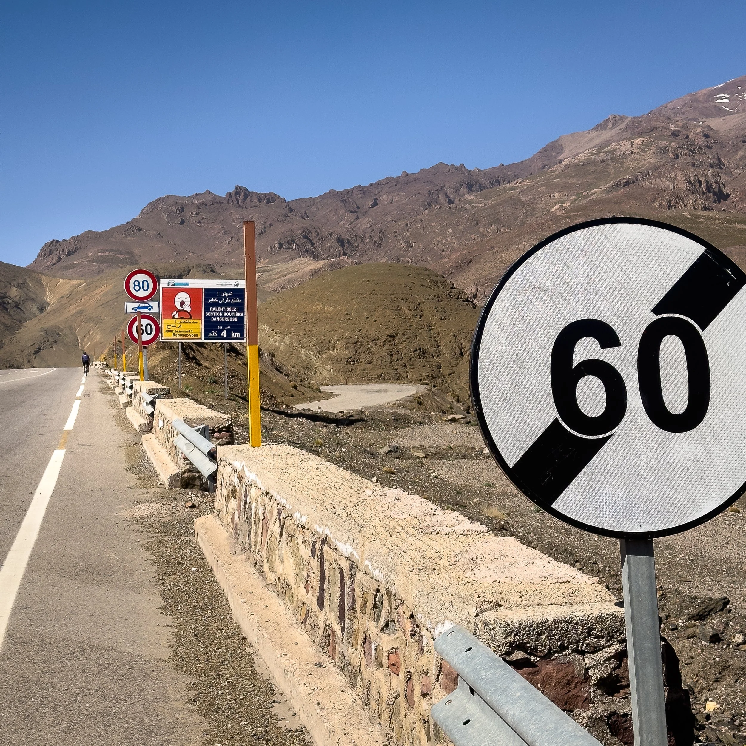 Mountain road with multiple speed limit signs, the largest sign showing a speed limit of 60, with other signs indicating 80 and 50, and a warning sign about a dangerous section of the route. Mountains in the background under a clear blue sky.