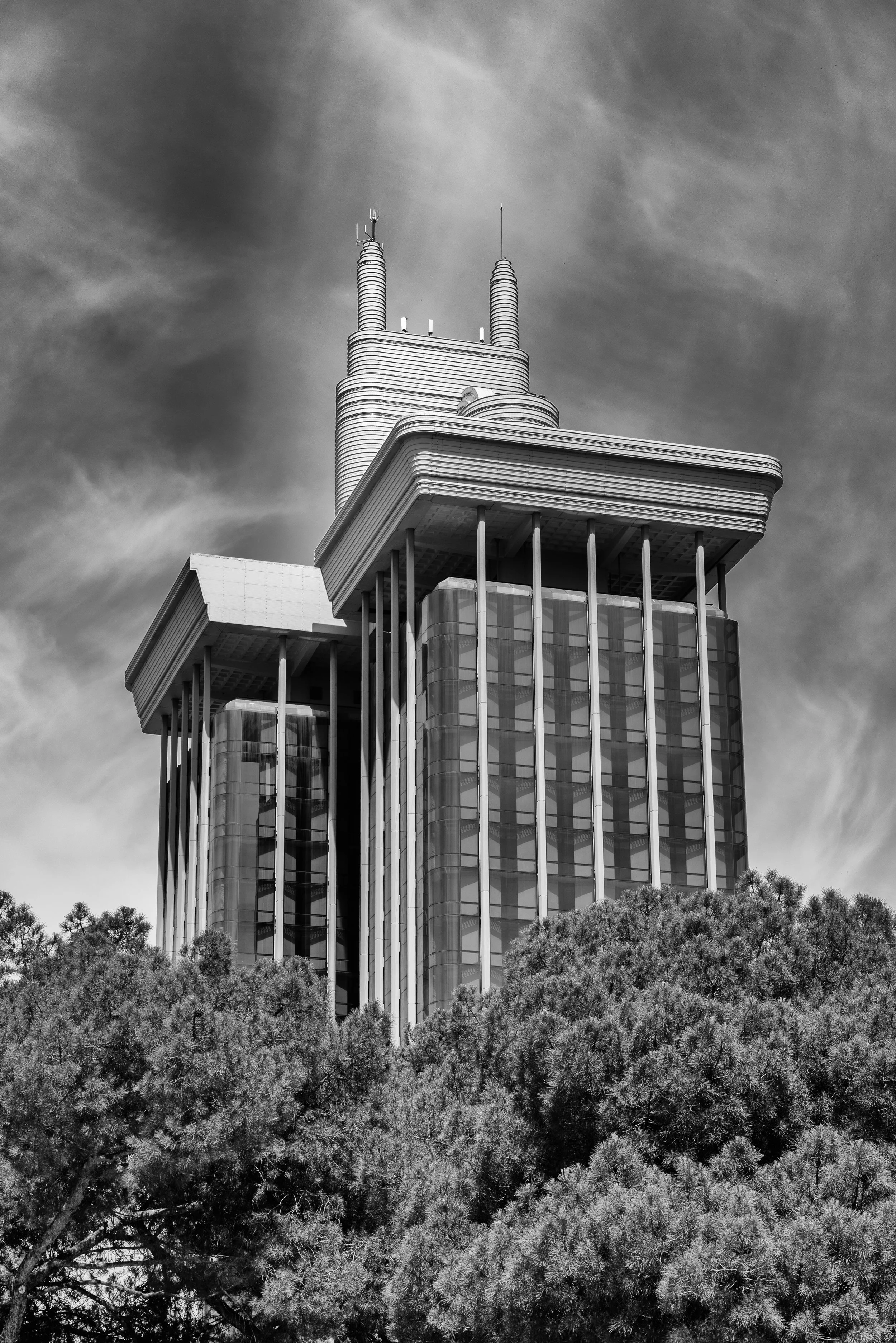Black and white photo of a modern skyscraper with trees at the bottom, clouds in the sky above.