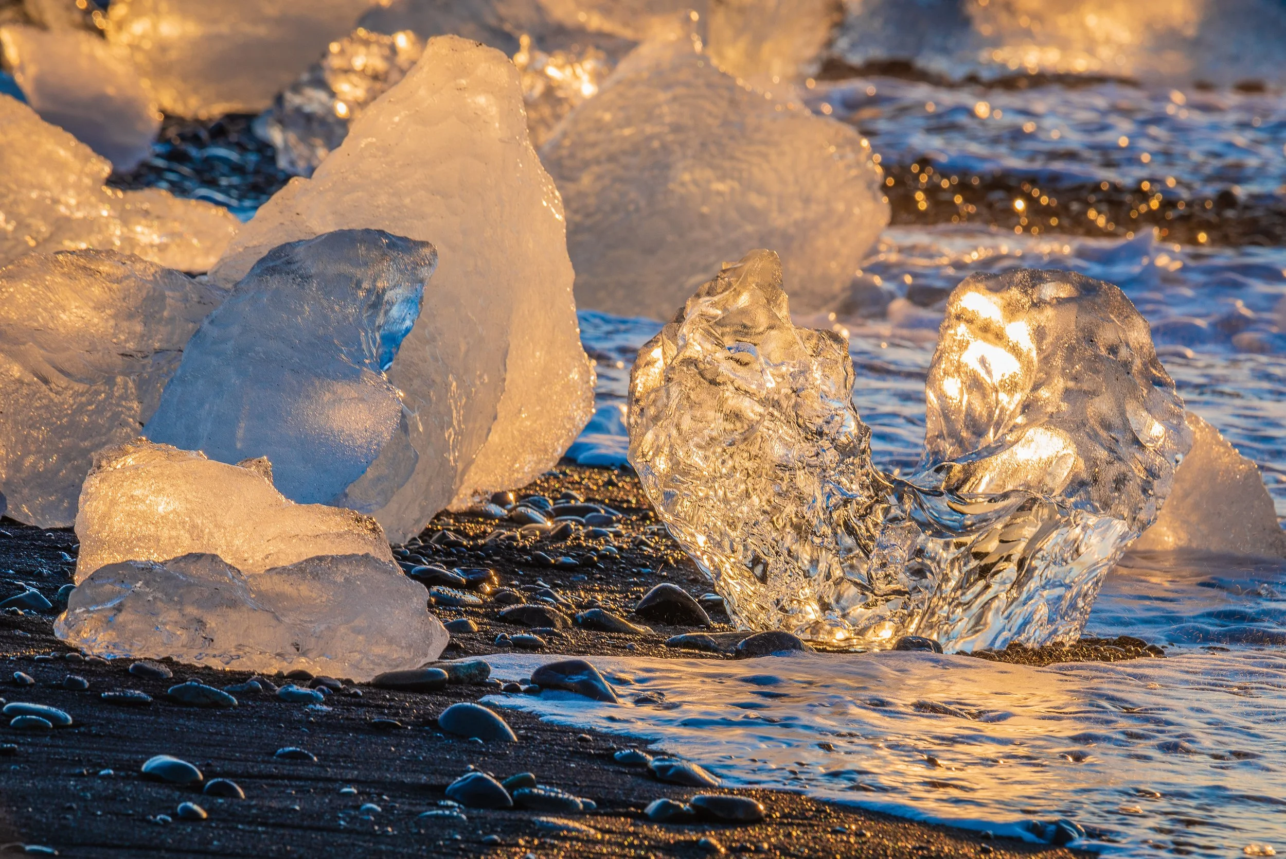 Ice chunks and pebbles on a beach with waves, illuminated by golden sunlight.