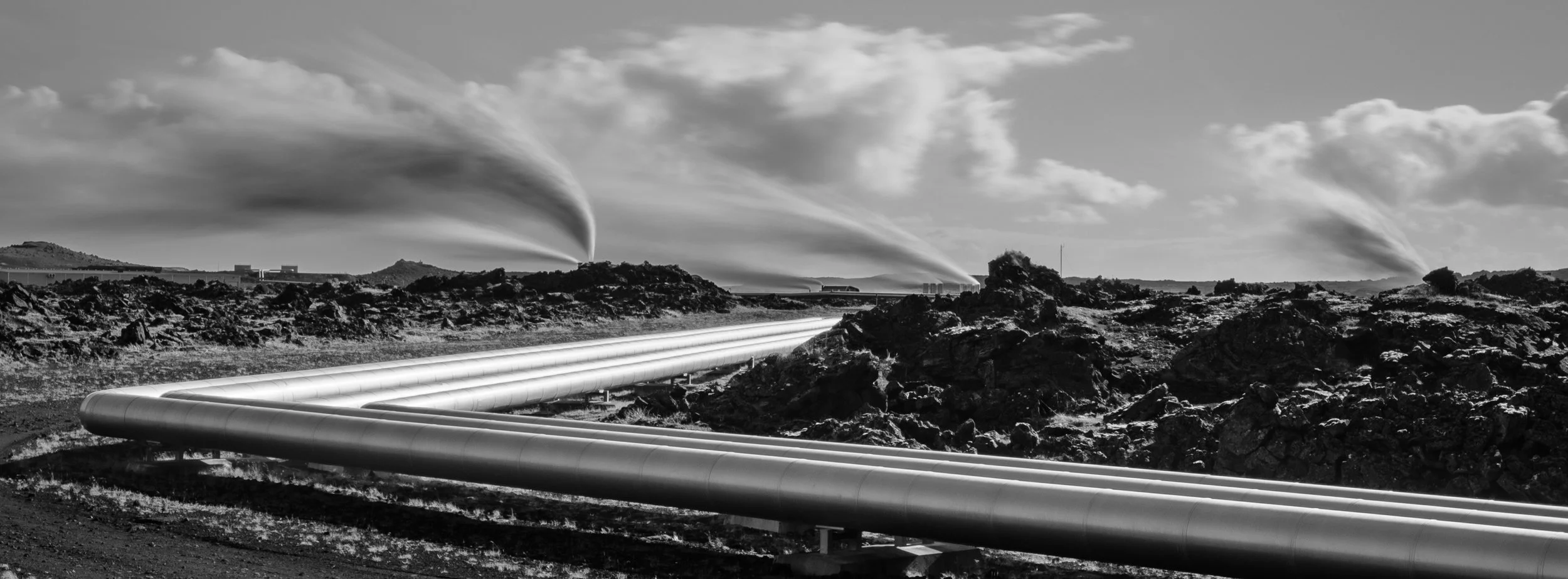 Black and white photo of industrial landscape with large pipelines running across rocky terrain, smoke or steam rising from distant cooling towers, and a cloudy sky.