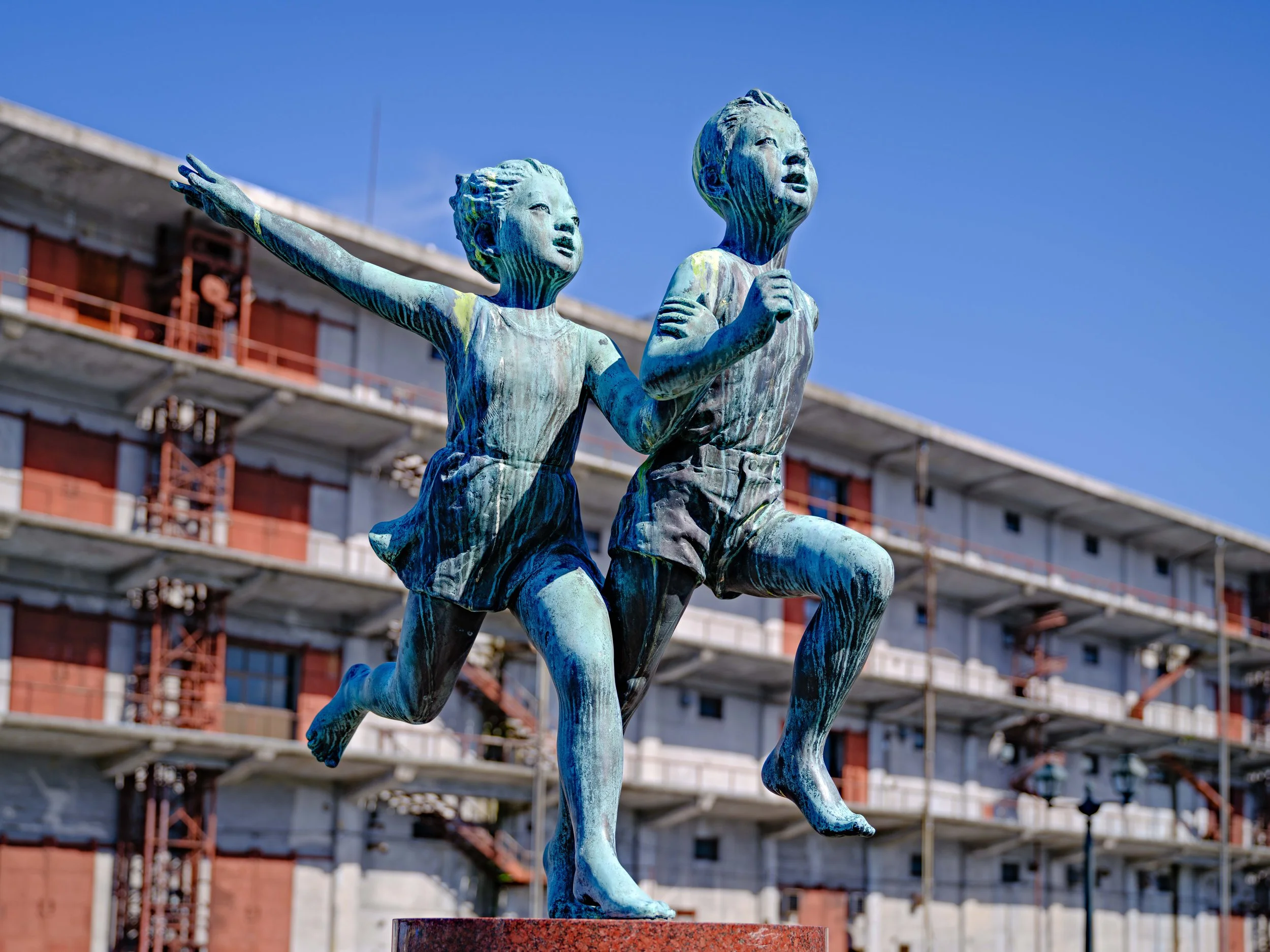 Bronze statue of two children running, with a girl in front and a boy behind, against a backdrop of a multi-story building under construction and a clear blue sky.