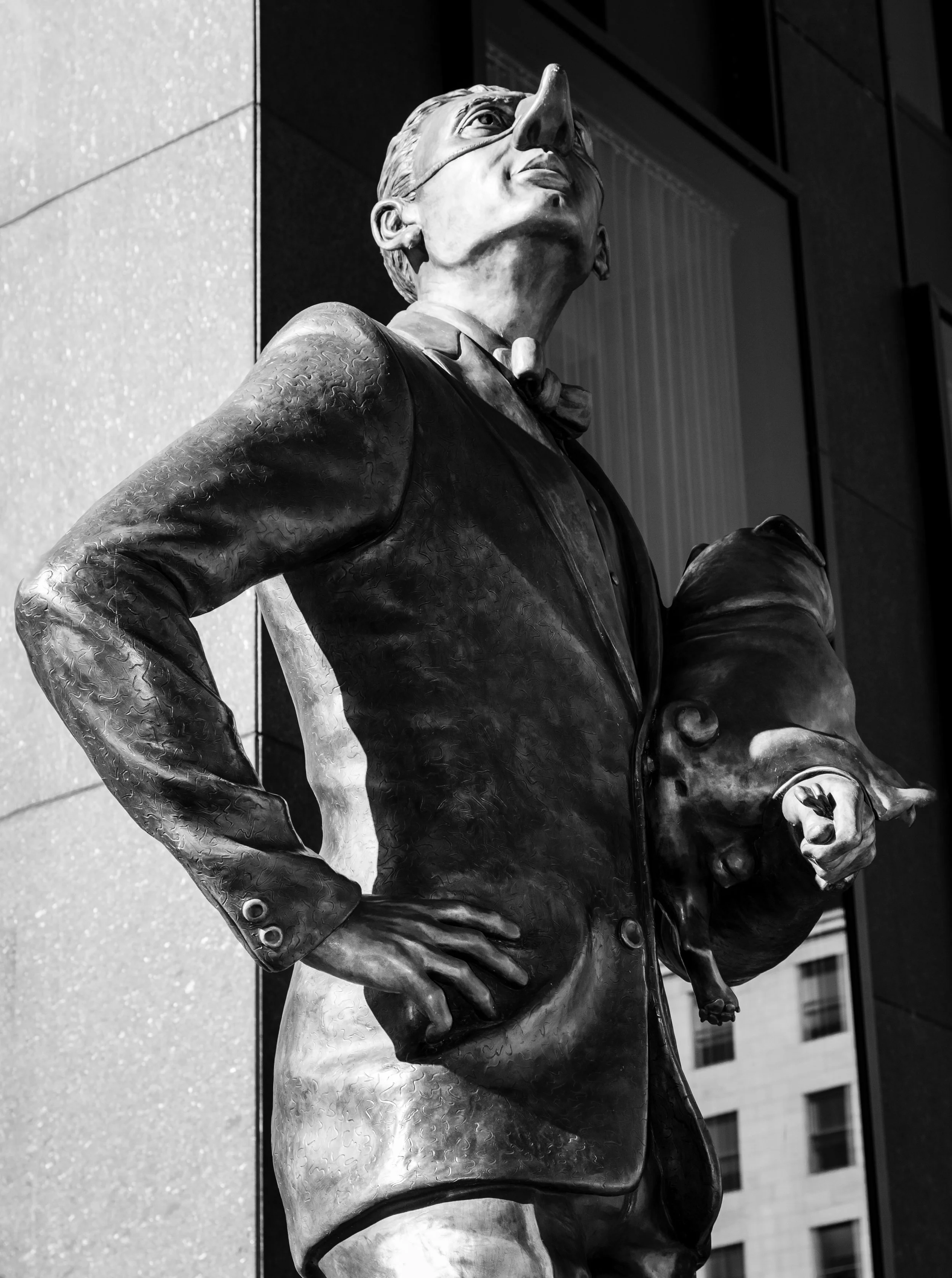 Black and white photo of a statue of a man in a suit holding a pig under his arm, standing against a modern building background.