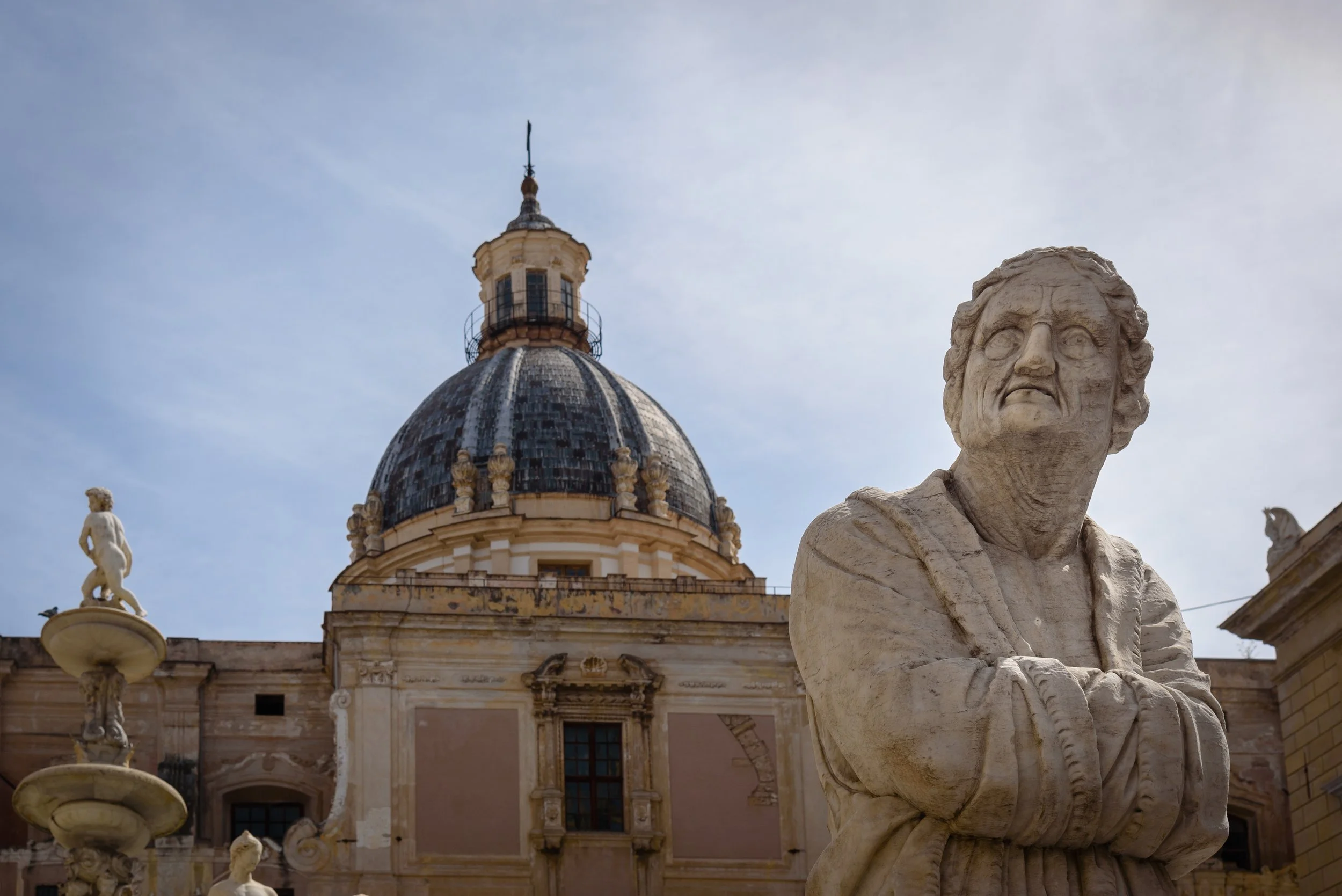 Marble statue of a man with crossed arms in front of historic European building with dome and statues.