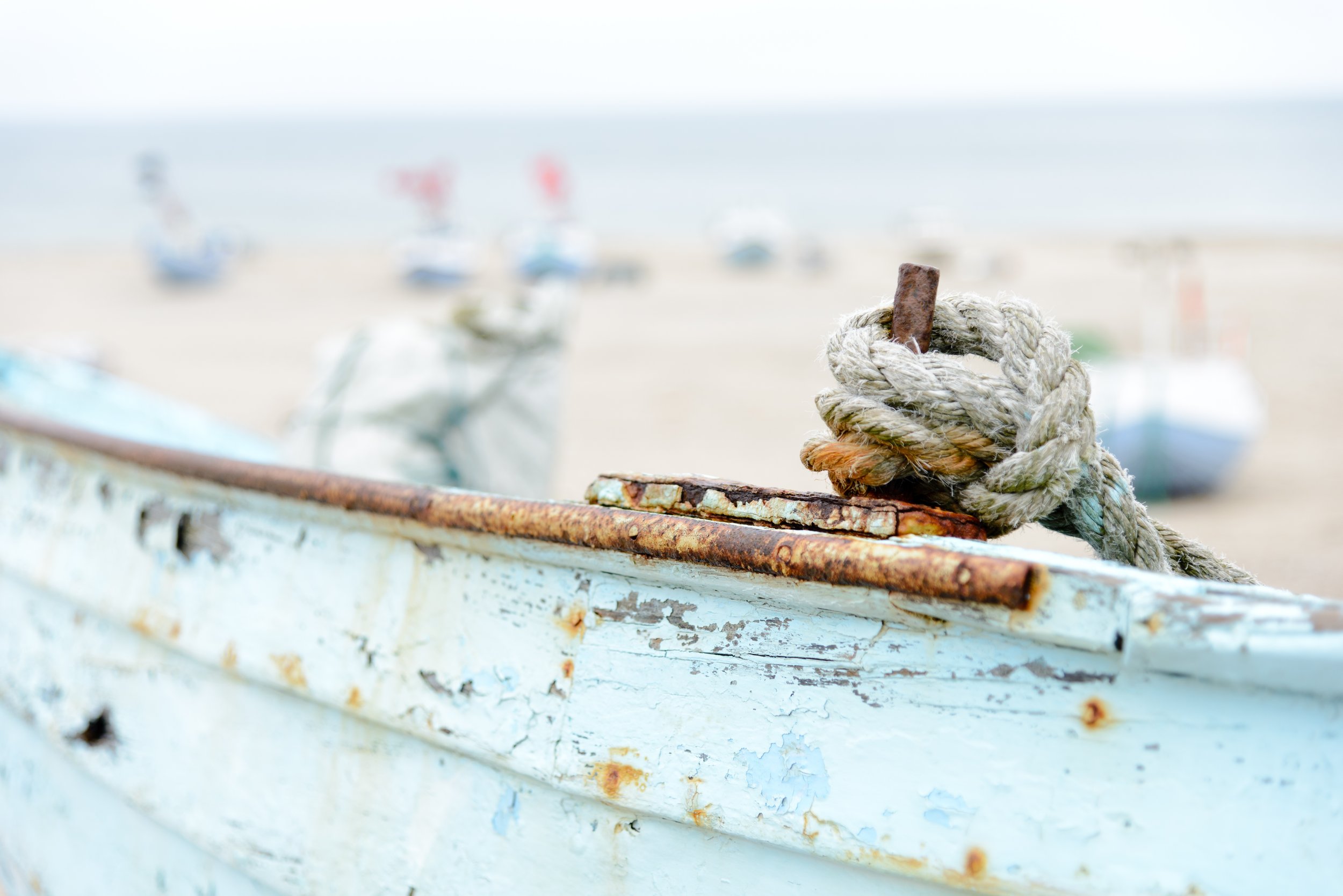 Close-up of a rusted boat with a weathered rope tied around a metal hook, on a sandy beach with boats and the ocean in the background