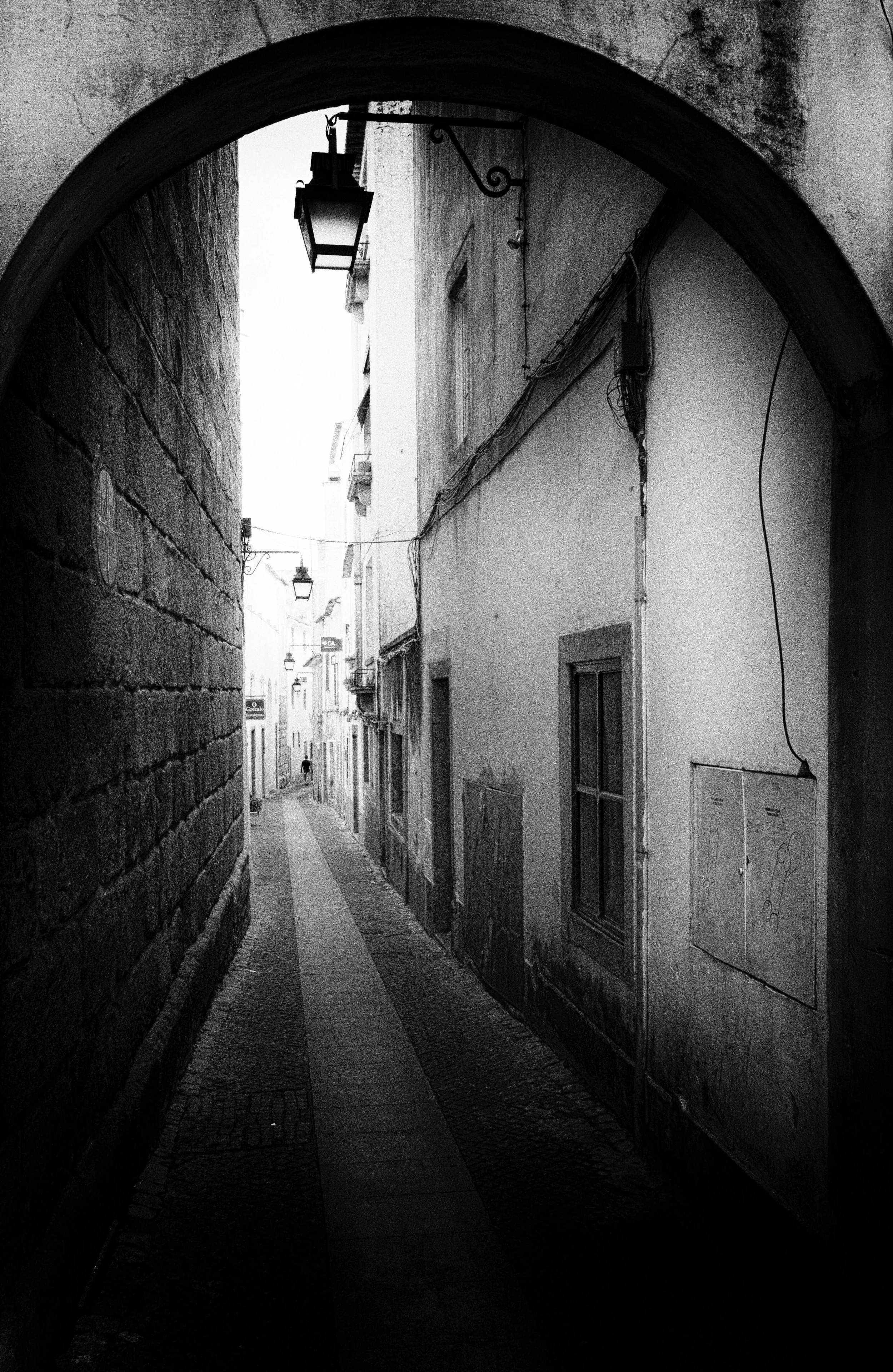 Black and white photo of a narrow alleyway seen through an archway, with aged buildings, windows, and hanging street lamps.