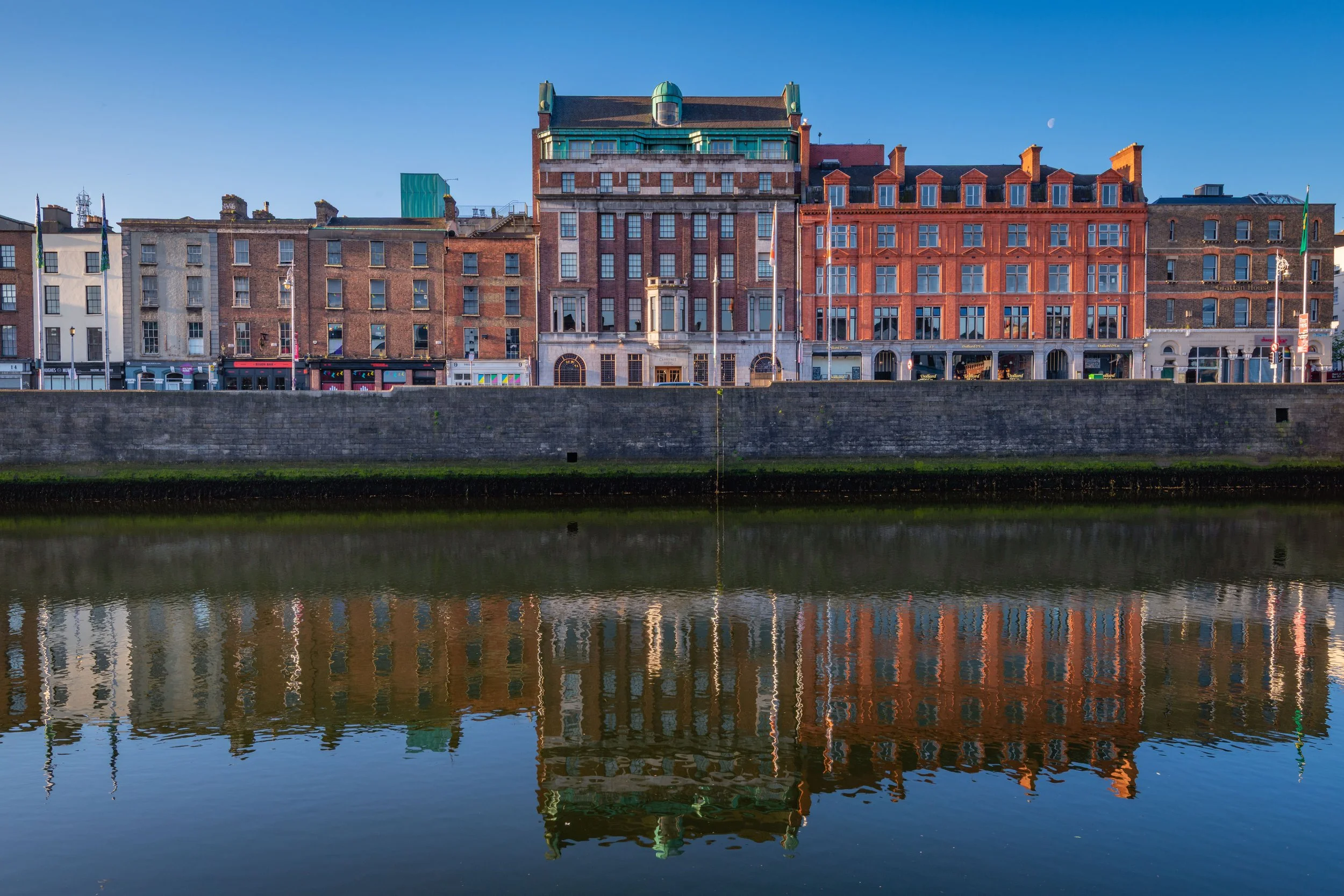 A row of historic buildings along a riverbank, with their reflections visible in the water, under a clear blue sky.