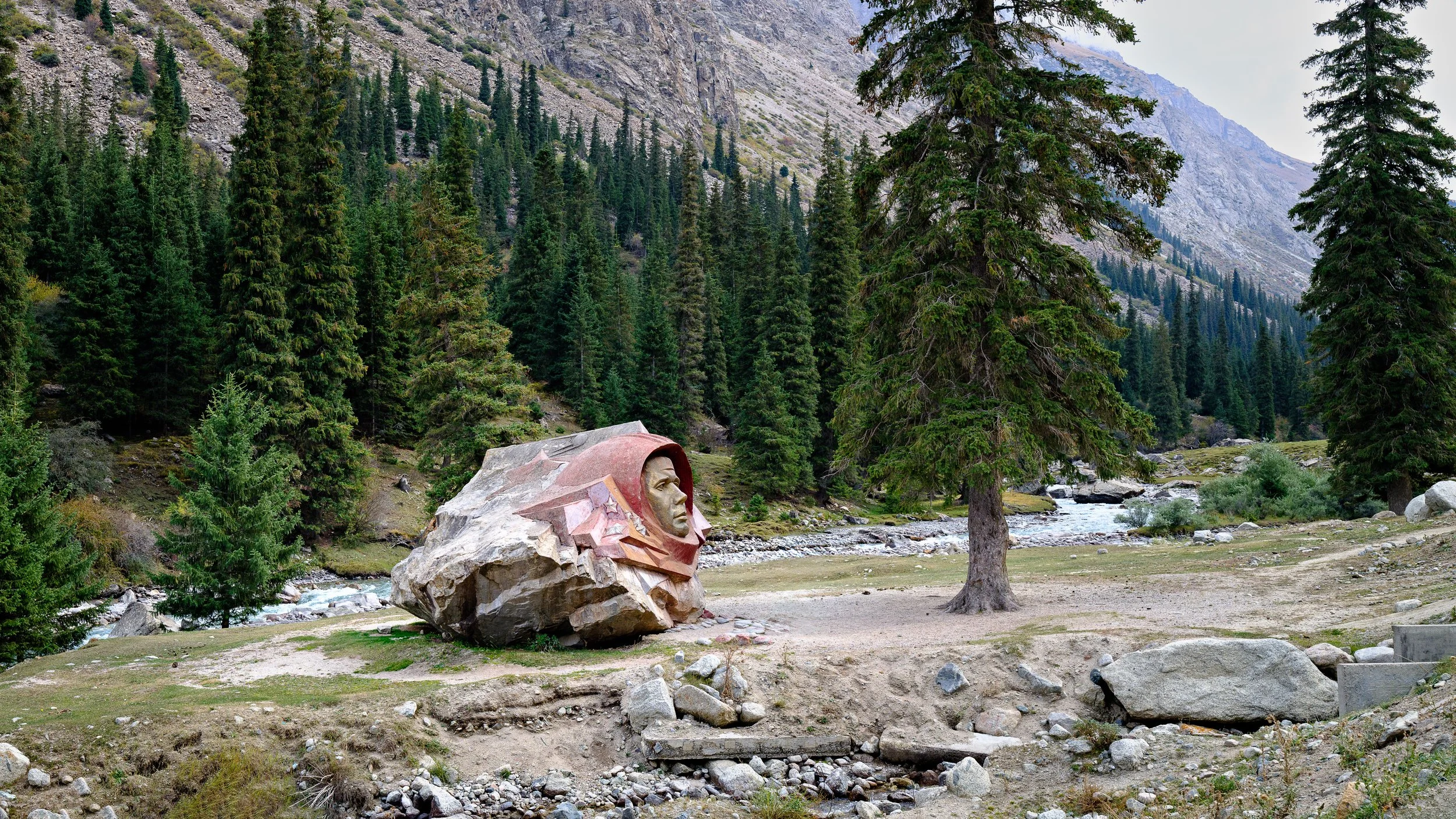 A memorial sculpture of a person's face embedded in a large rock, surrounded by pine trees in a mountainous landscape with a river in the background.