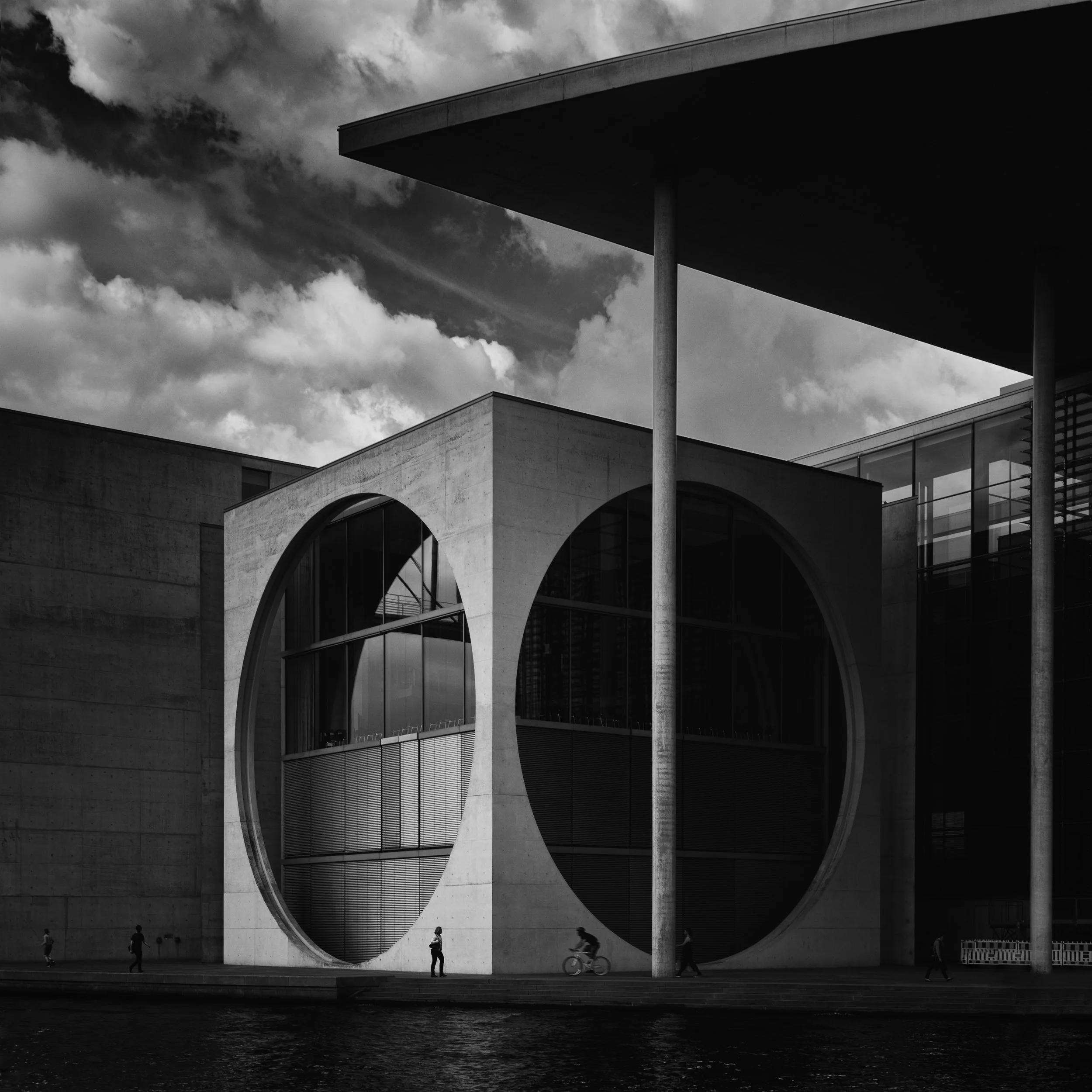 Black and white photo of modern building with large circular windows and a overhanging roof, with people walking and cycling nearby, under cloudy sky.
