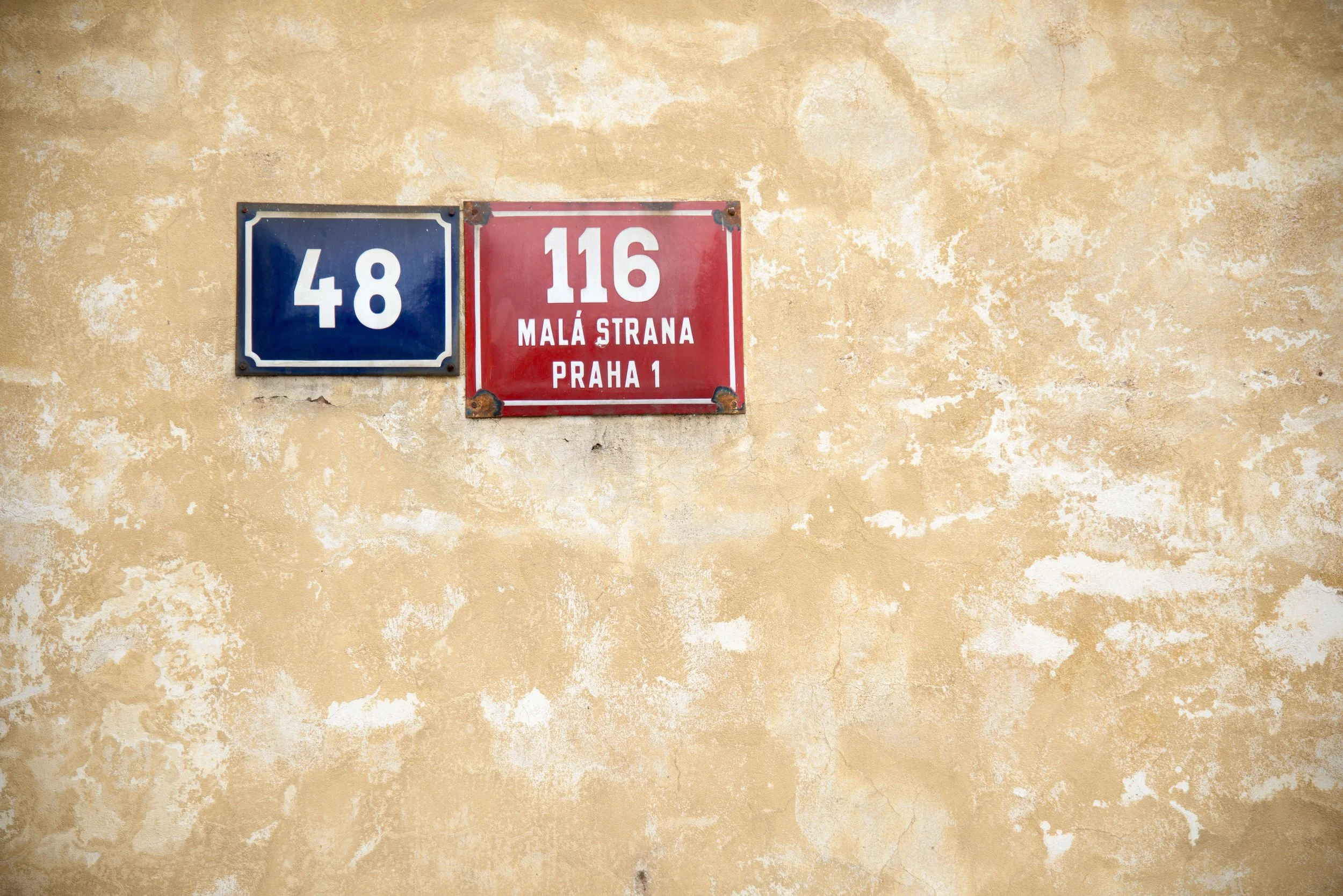 Two street signs mounted on a weathered yellow wall. The blue sign displays the number 48, and the red sign shows the number 116 with the text 'Malá Strana Praha 1'.
