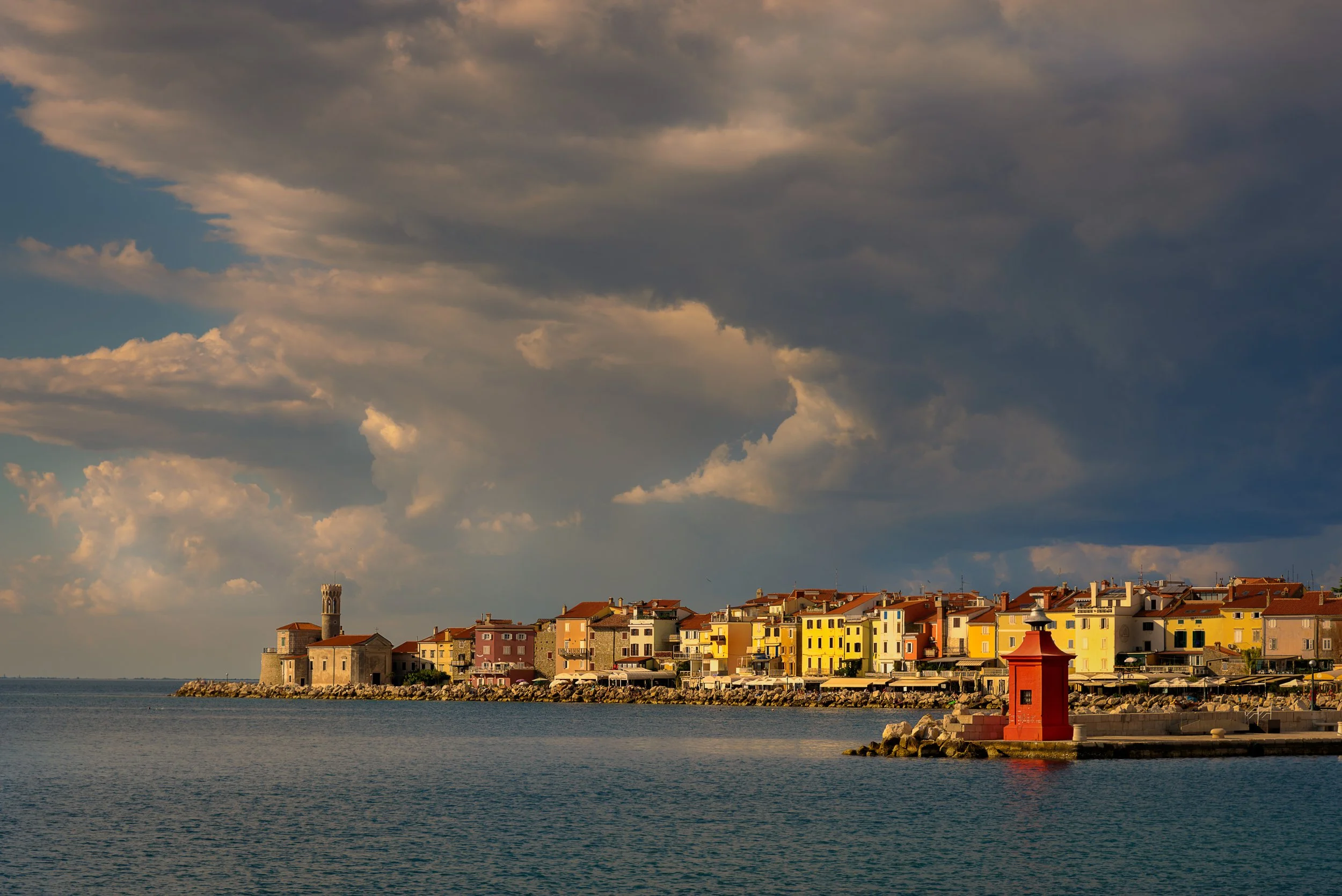 A coastal town with colorful buildings along the shoreline, a small red lighthouse on a jetty, under a cloudy sky at sunset.