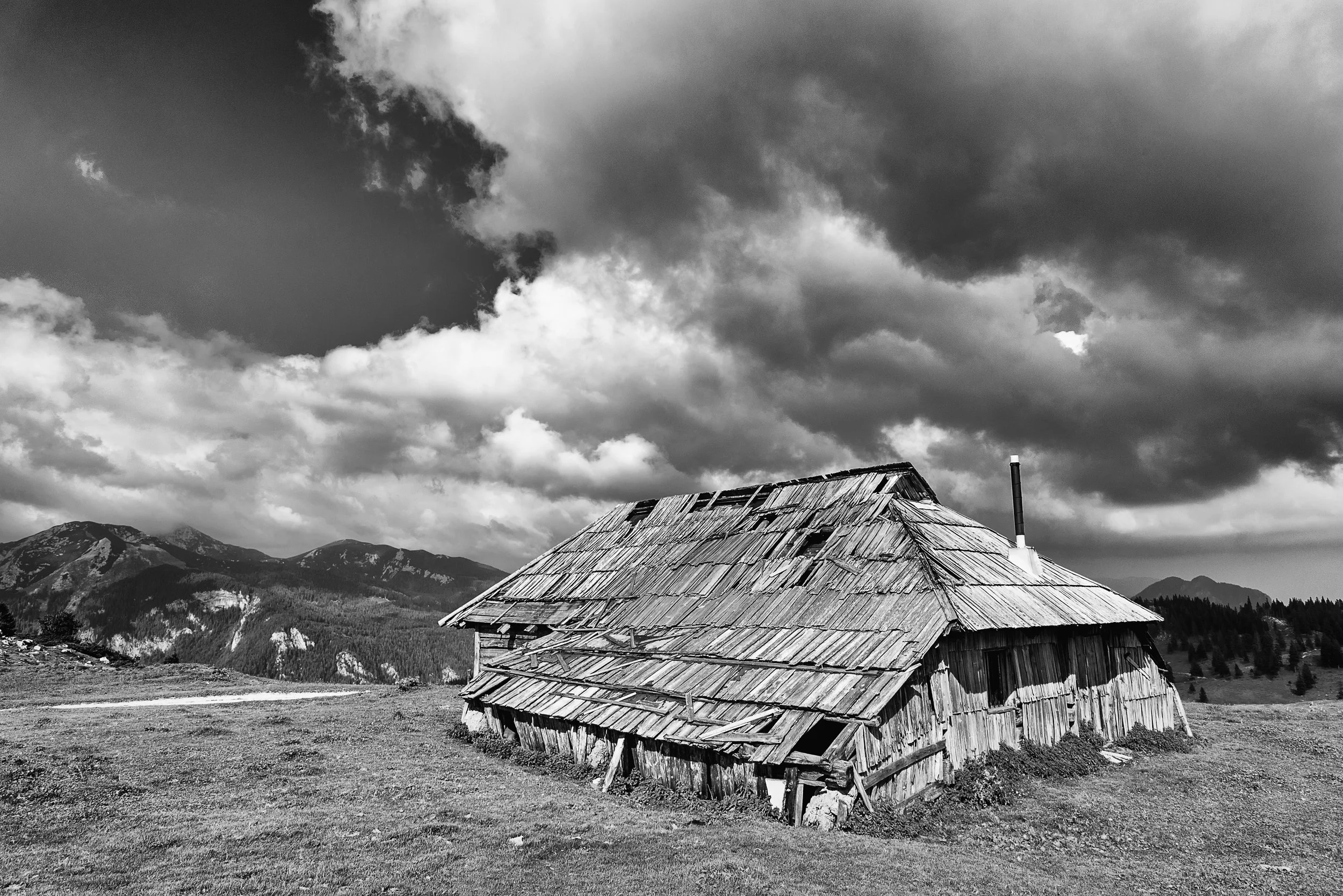 An old, dilapidated wooden house sitting in an open field with mountains in the background under a cloudy sky.