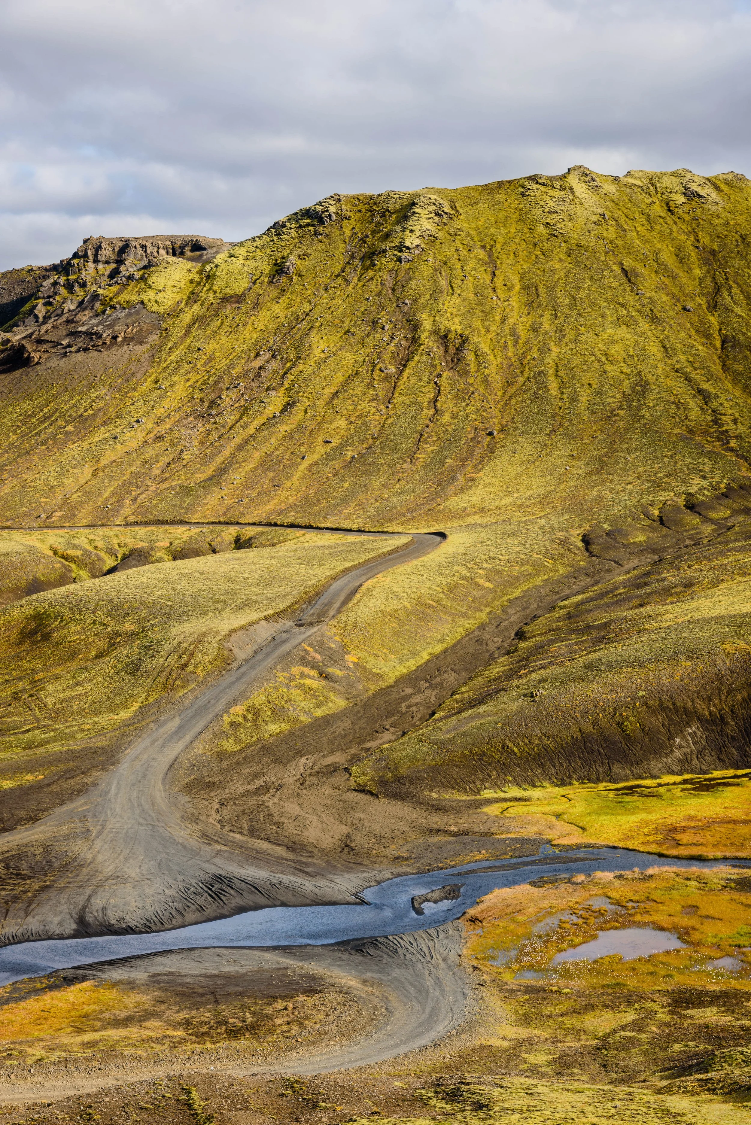 A winding dirt road runs through a mountainous landscape covered in green moss, with a small stream near the bottom, under a cloudy sky.