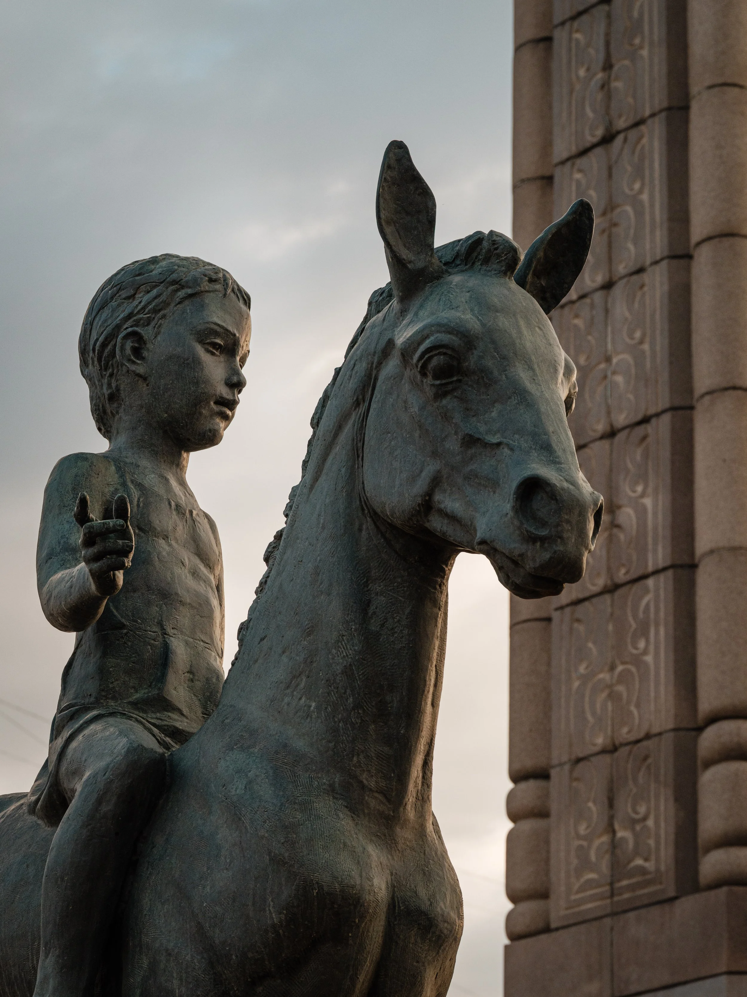 Bronze sculpture of a young boy riding a horse near a stone arch.