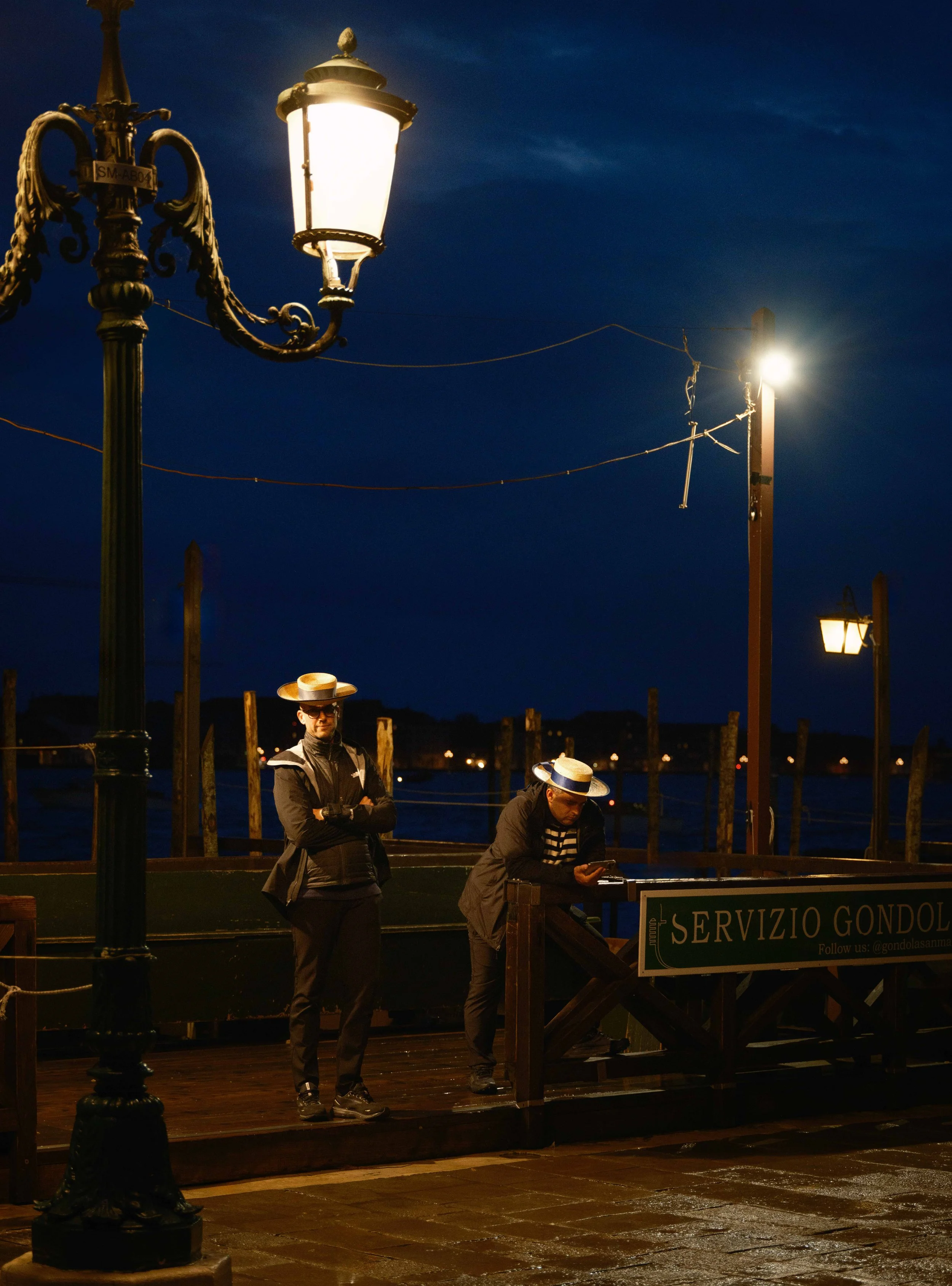 Nighttime scene at a dock with two men wearing black jackets and striped shirts, both wearing wide-brimmed hats. One man stands with arms crossed, the other appears to be looking at a device. Old-fashioned street lamps illuminate the scene, and there