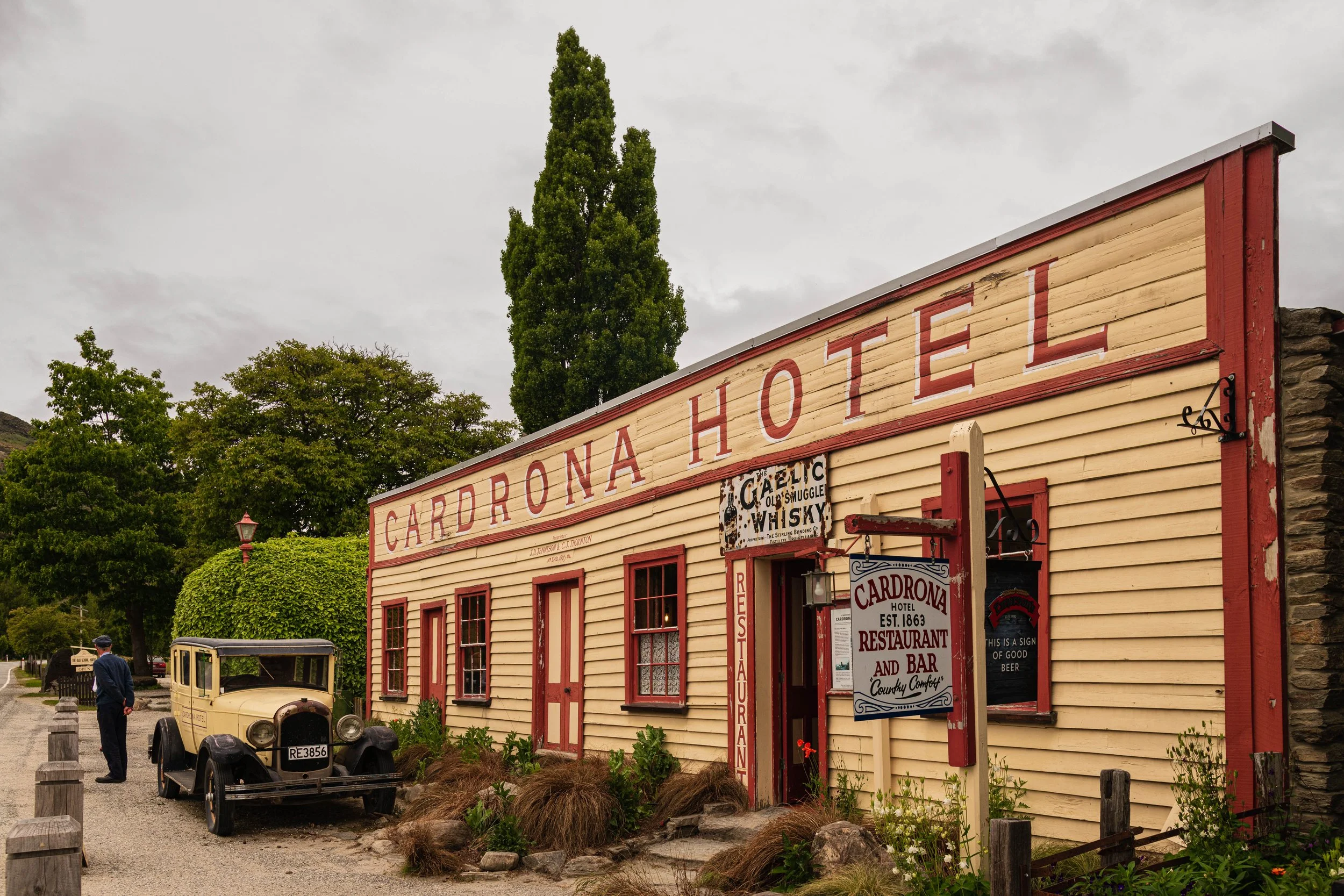 Historic building labeled 'Cardrona Hotel' with vintage car parked outside and a man in a uniform nearby. The building has a rustic look with yellow wood siding and red trim, and signs for the hotel, restaurant, and bar. There are trees and overcast 