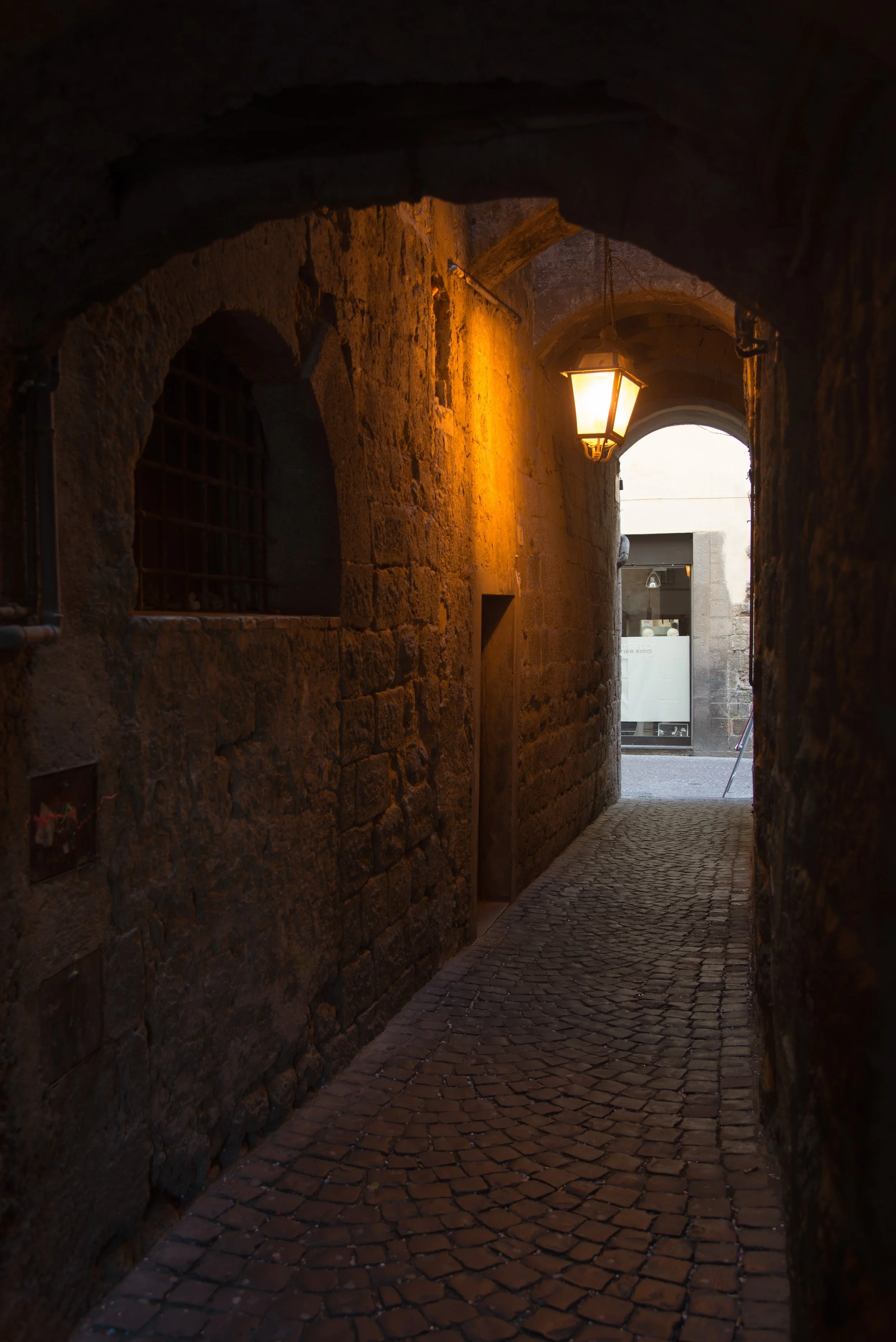A narrow, cobblestone alleyway with stone walls, an arched entrance, and a hanging lantern illuminating the scene