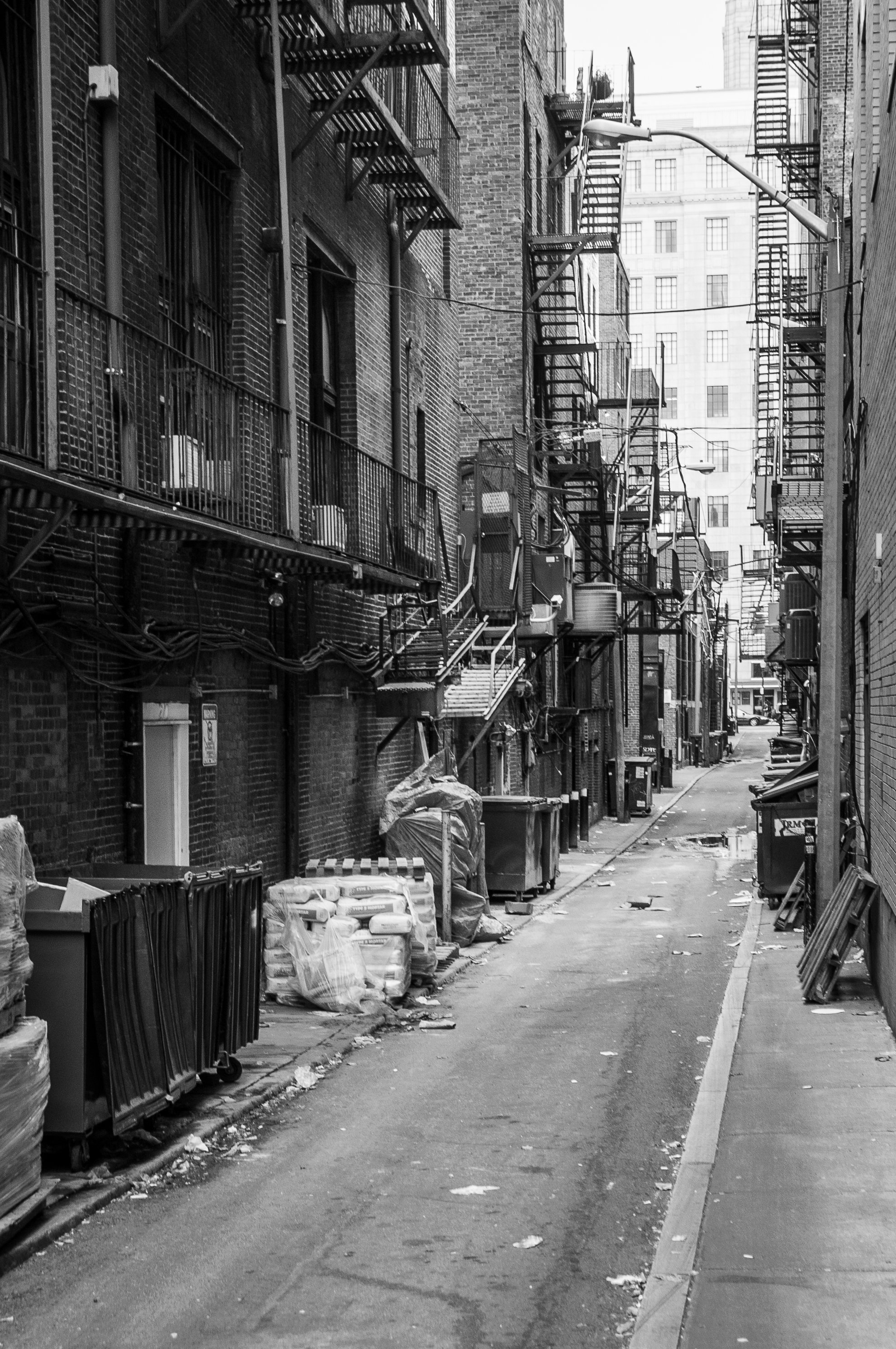 Alleyway between old brick buildings with fire escapes, trash bins, wrapped pallets, and scattered debris in black and white.