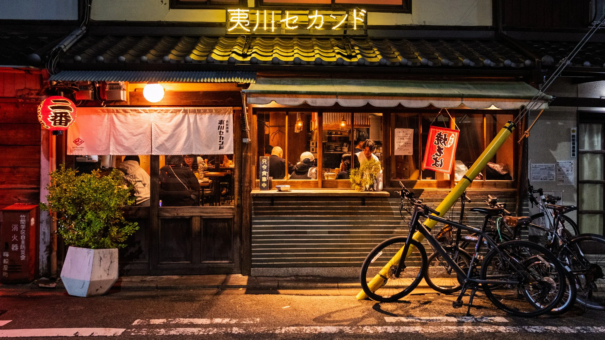 Nighttime street scene outside a Japanese restaurant with bicycles parked outside. The restaurant has warm lighting, a wooden exterior, and Japanese signs, with people dining inside.