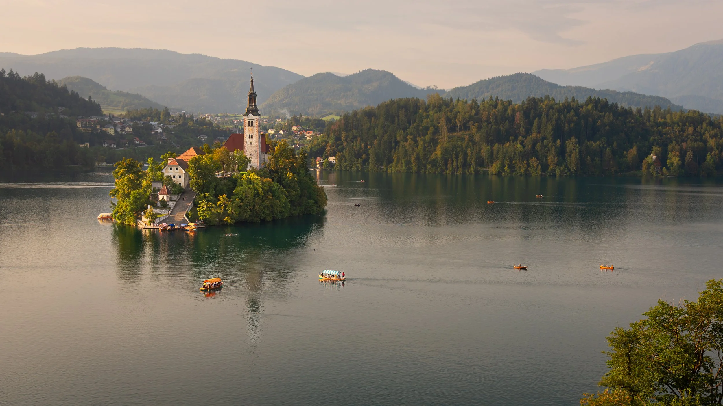 A scenic view of Lake Bled in Slovenia with an island featuring a church and buildings, surrounded by lush green forests and mountains in the background, with several boats on the water.