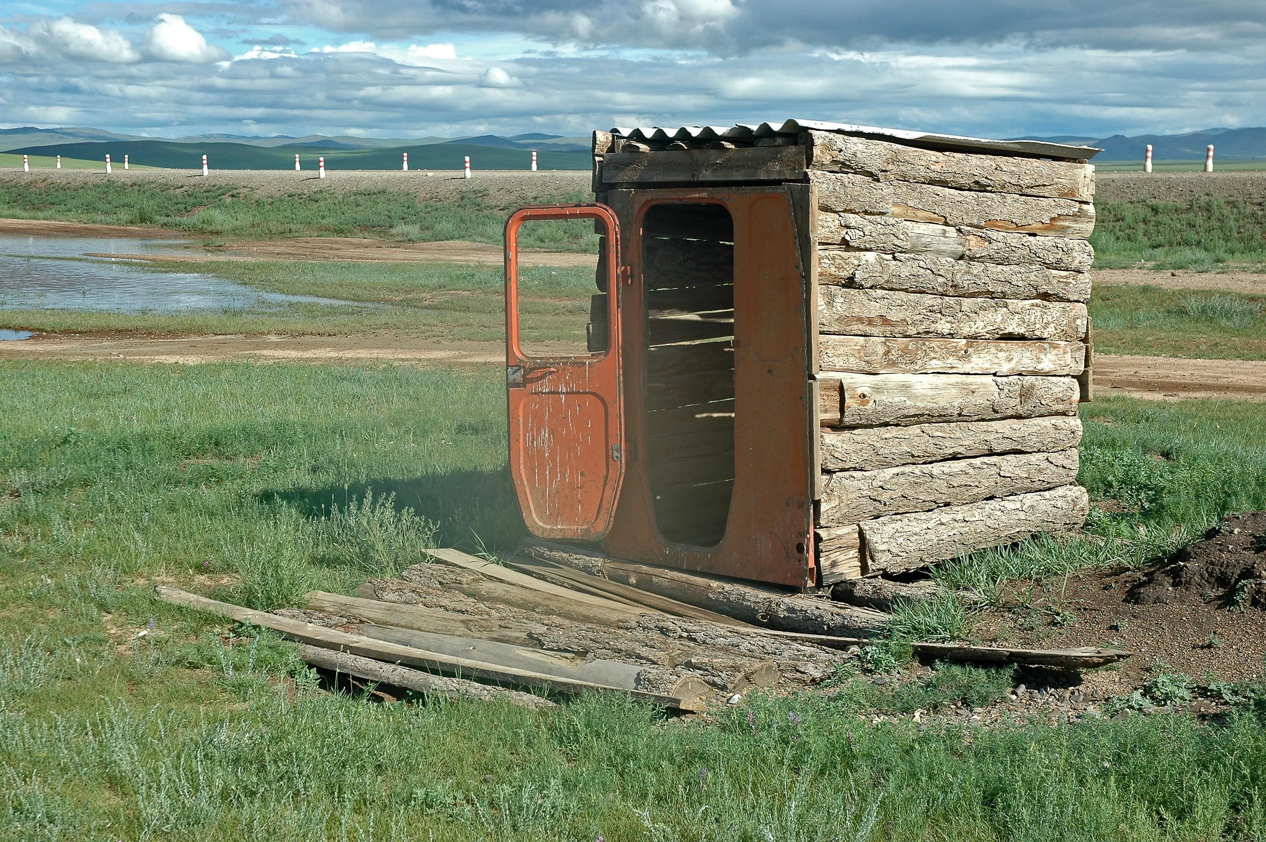 A rusty old truck door propped up against a small wooden structure made of logs in a grassy open field.