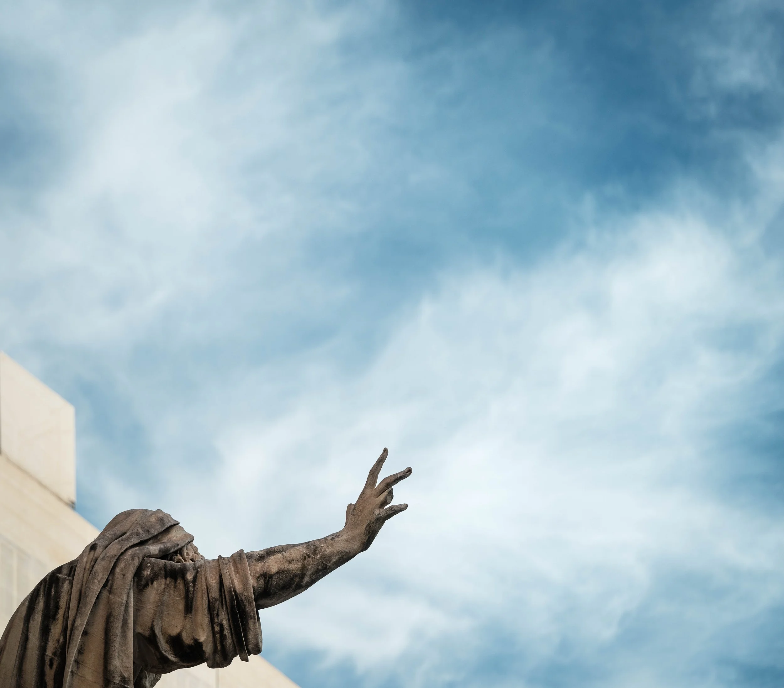 Close-up of a sculpture of an elderly woman with her arm extended and finger pointing, set against a blue sky with scattered clouds.