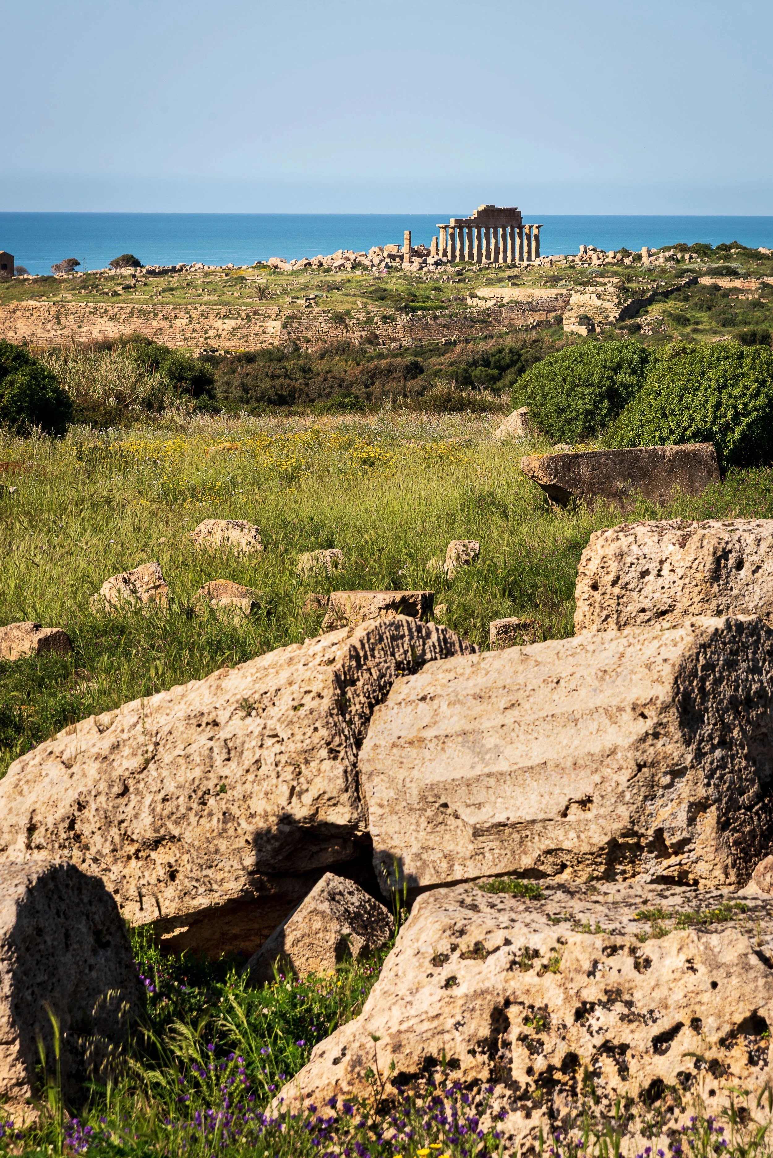 Ancient ruins with large boulders and green grass in the foreground, a partially preserved Greek temple with columns in the middle ground, and the sea in the background.