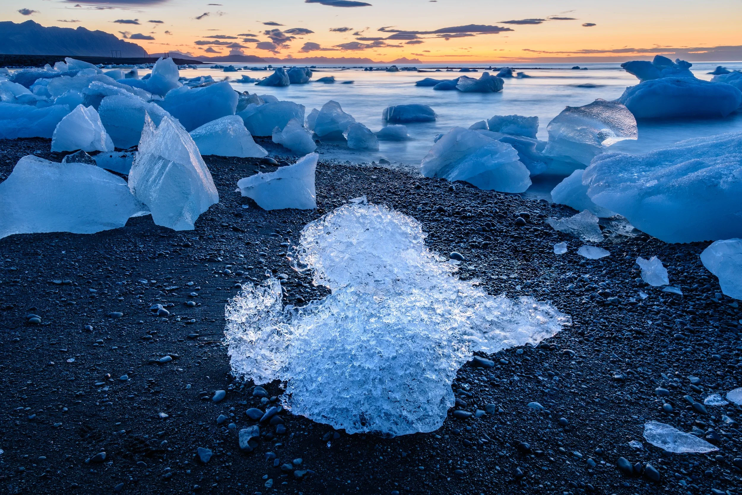 Ice chunks on a black pebble beach at sunset, with the ocean and a distant mountain in the background.