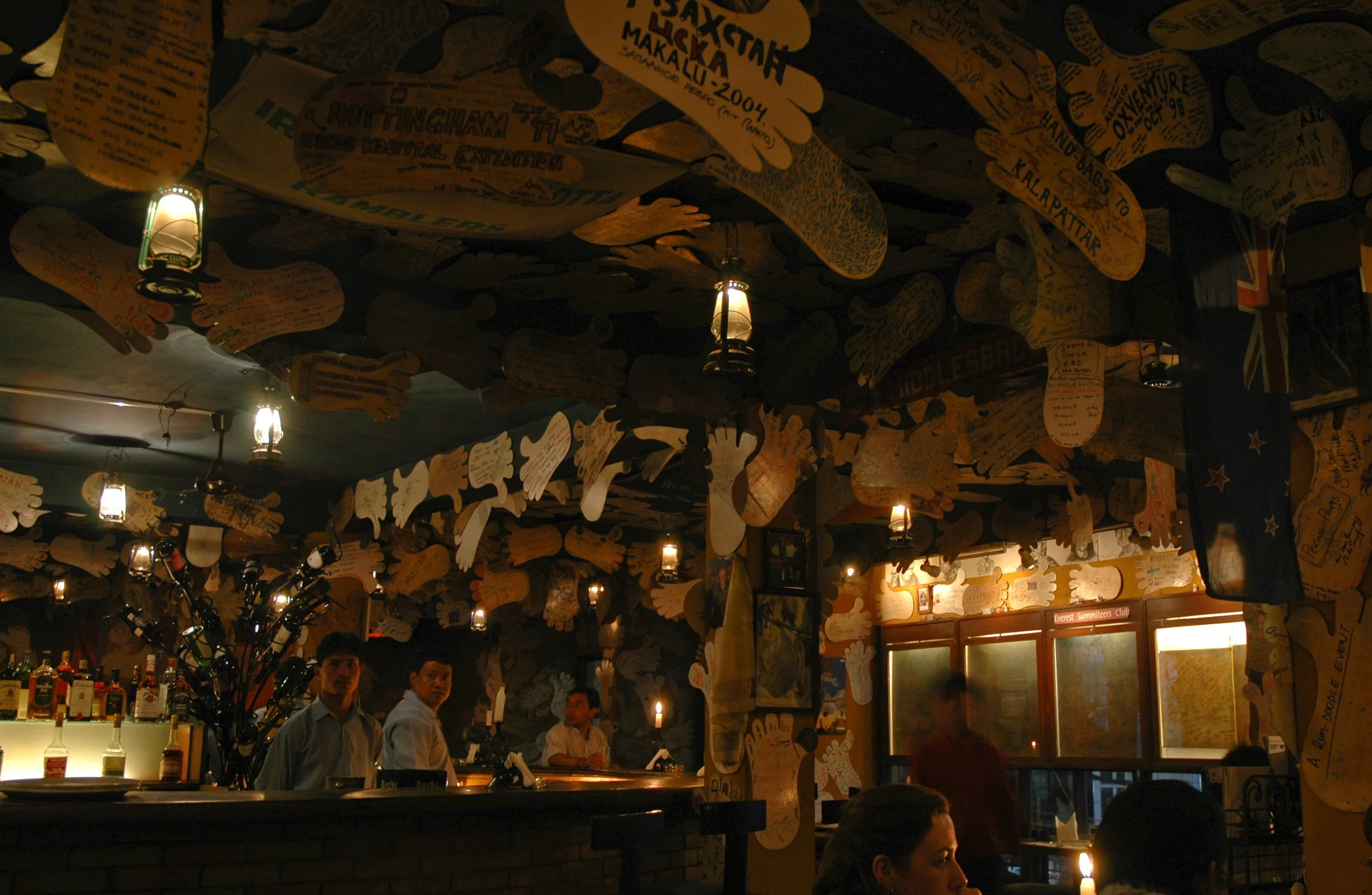 Interior of a dimly lit bar or restaurant with walls and ceiling decorated with numerous paper cutouts in the shape of hands, and small hanging lantern-style lights. Several people are visible, including bartenders and patrons.