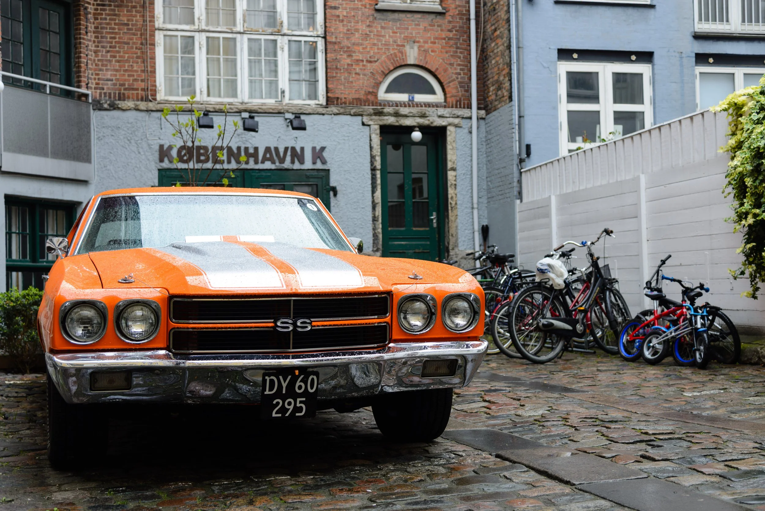 An orange vintage Chevrolet Camaro SS parked on a cobblestone street with bicycles in the background, and buildings with a sign that says 'KØBENHAVN K' behind it.