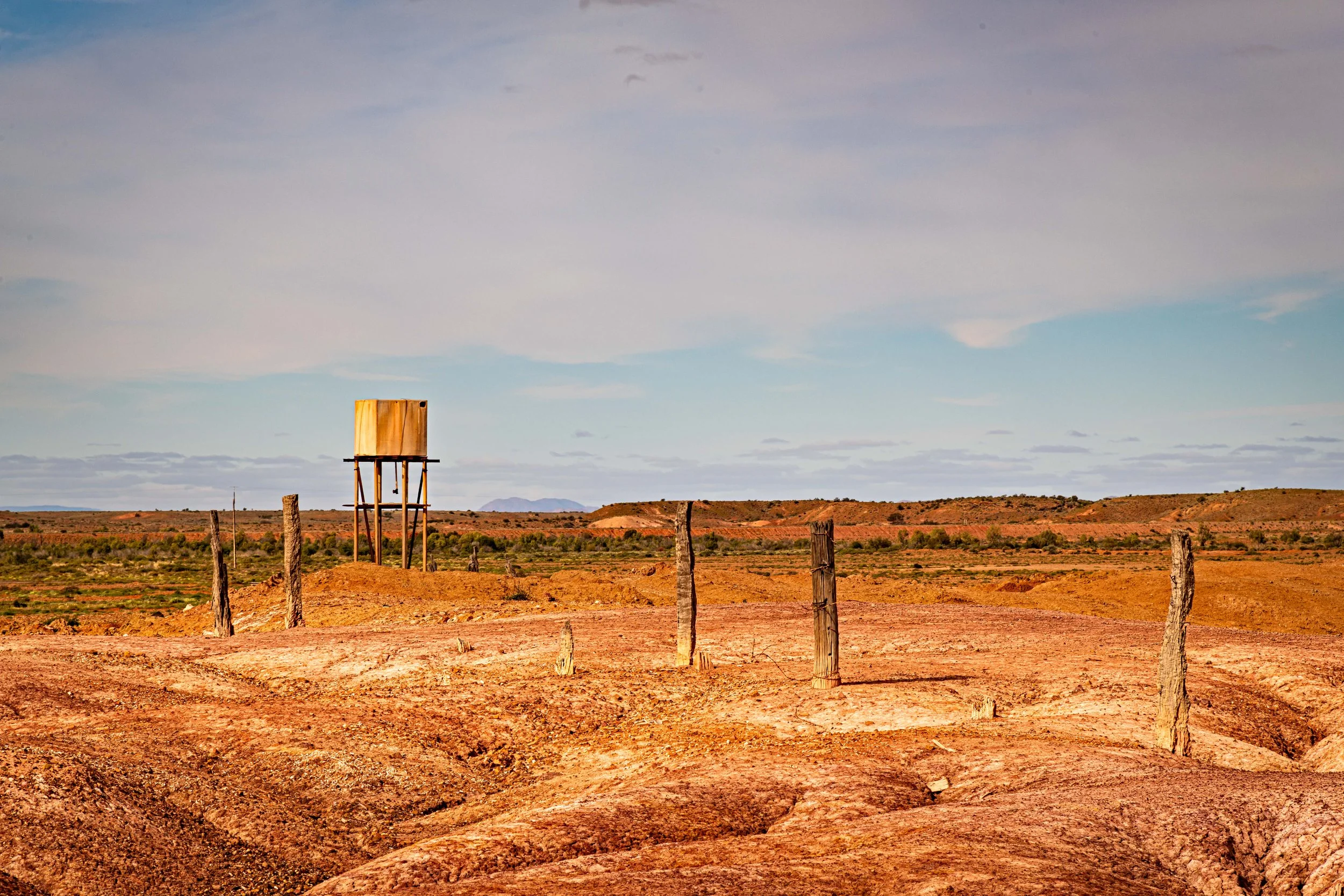 A barren desert landscape with dirt roads, weathered wooden fence posts, and an empty wooden water tower in the distance under a partly cloudy sky.