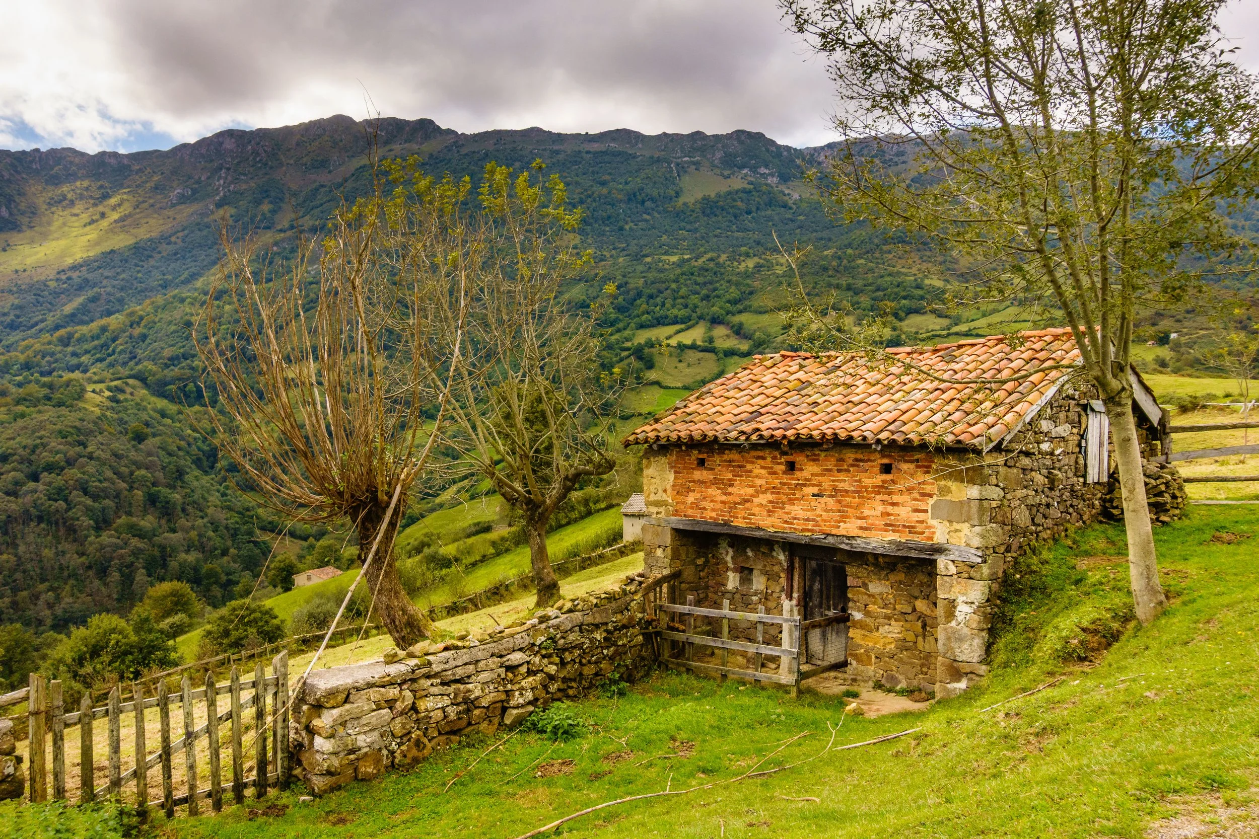 A small stone and brick cottage on a grassy hillside with leafless trees and mountains in the background.