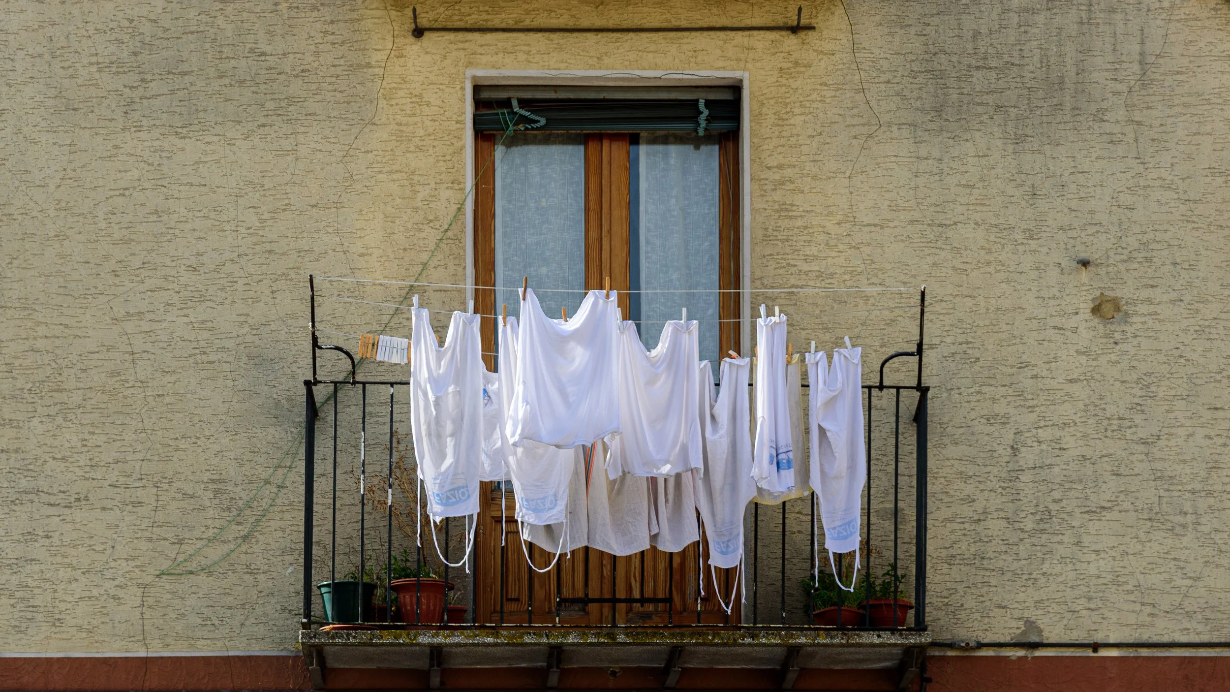 Clothes hanging on a clothesline on a small balcony outside a window, with potted plants below, against a yellow textured wall.