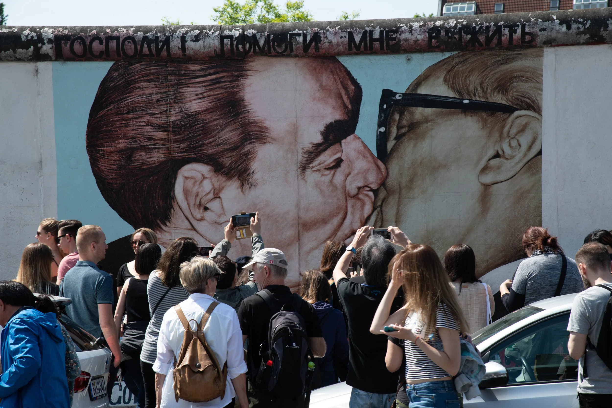 A large mural painting of two men kissing, one of whom is wearing glasses, with a crowd of people taking pictures in front of it.