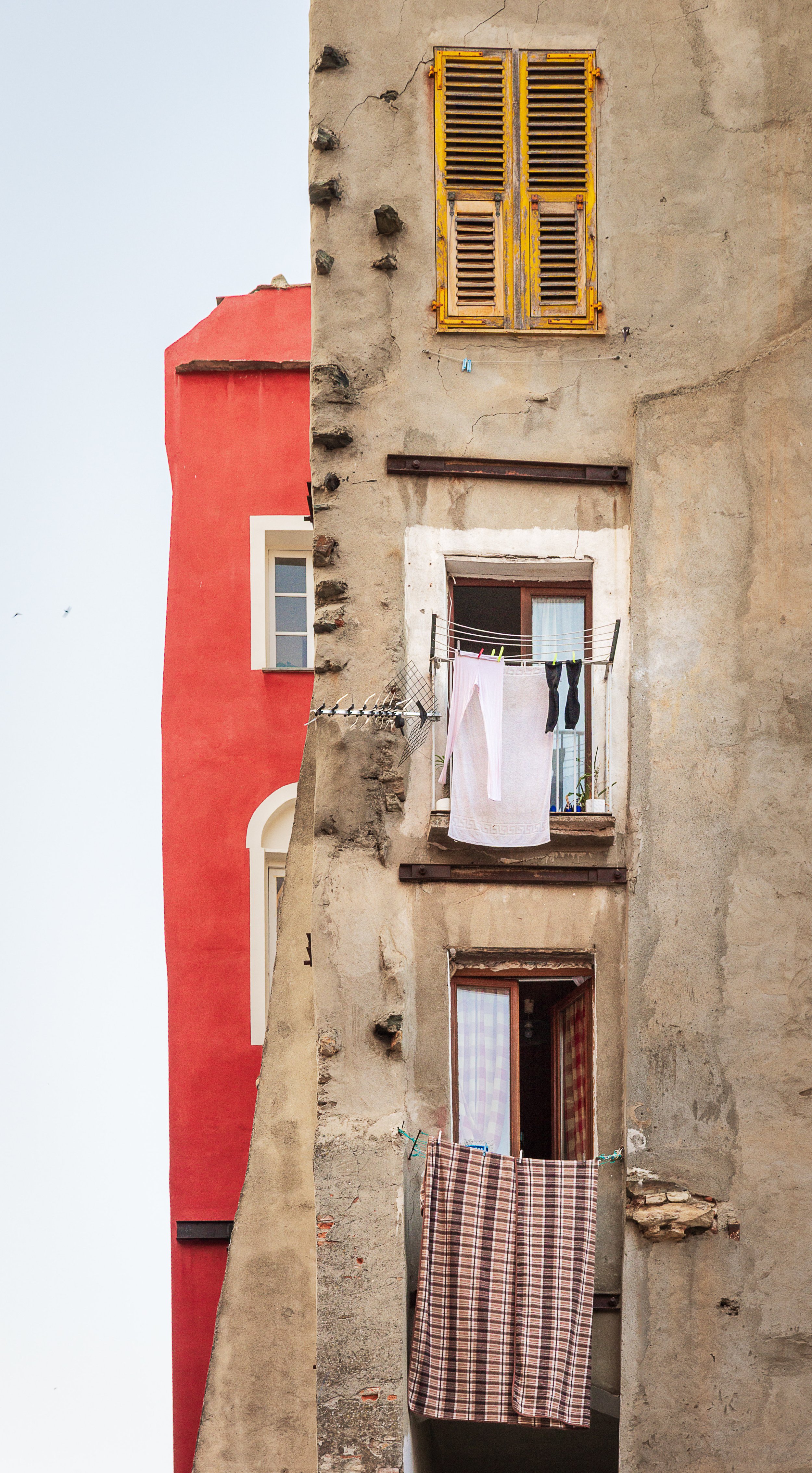 An old, weathered building with laundry hanging from a balcony, adjacent to a brightly painted red and beige building in an urban setting.