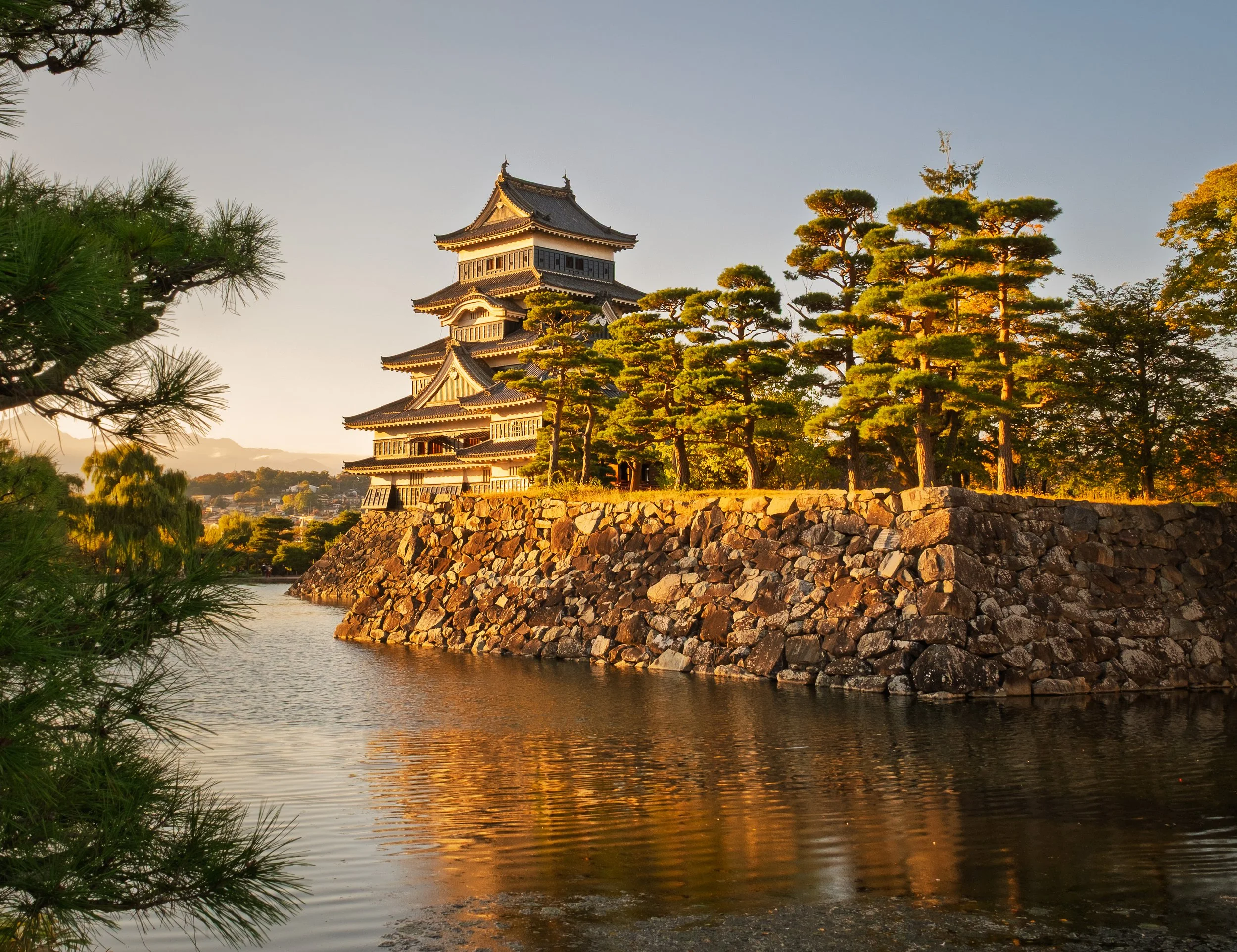 A traditional Japanese castle with multiple tiers and intricate roof designs, surrounded by trees and situated on a stone embankment beside a body of water during sunset.