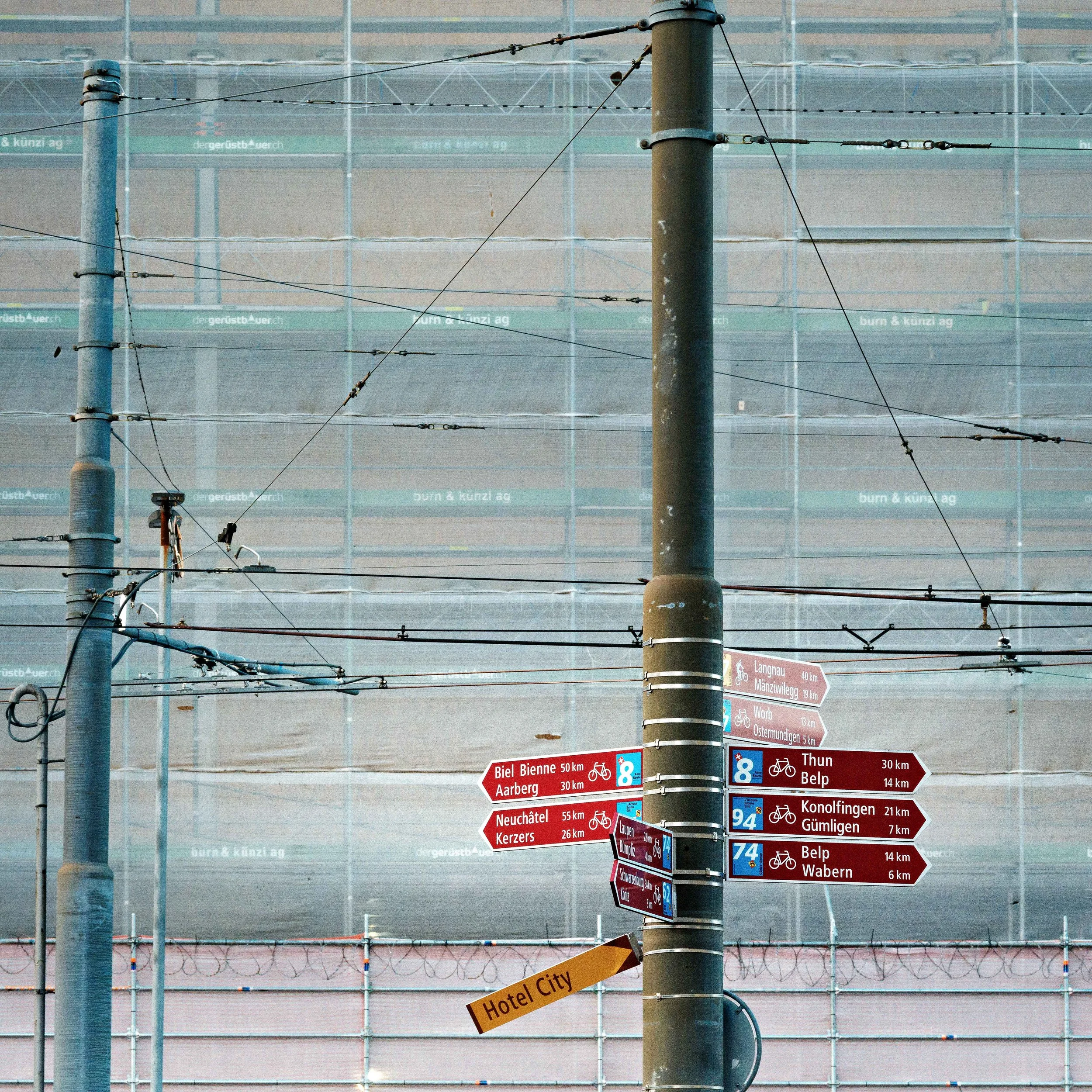 A street signpost with multiple directional signs pointing to various destinations and distances in kilometers, along with an orange sign labeled 'Hotel City,' set against a backdrop of a building covered in construction scaffolding with advertisemen