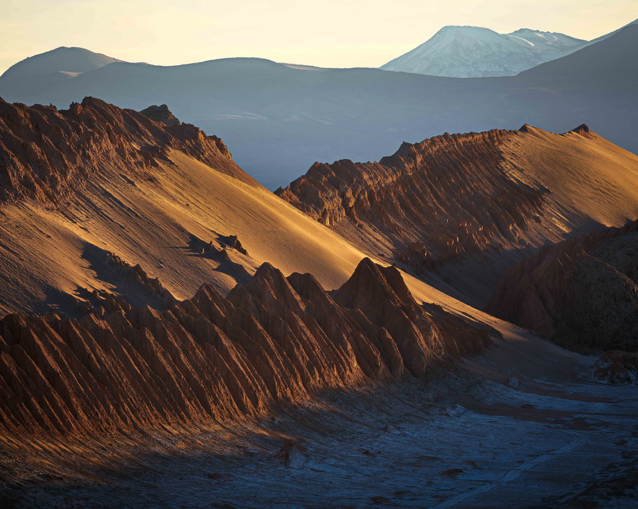 Mountain landscape with rugged reddish-brown peaks in the foreground and snow-capped mountains in the background, illuminated by sunset or sunrise.