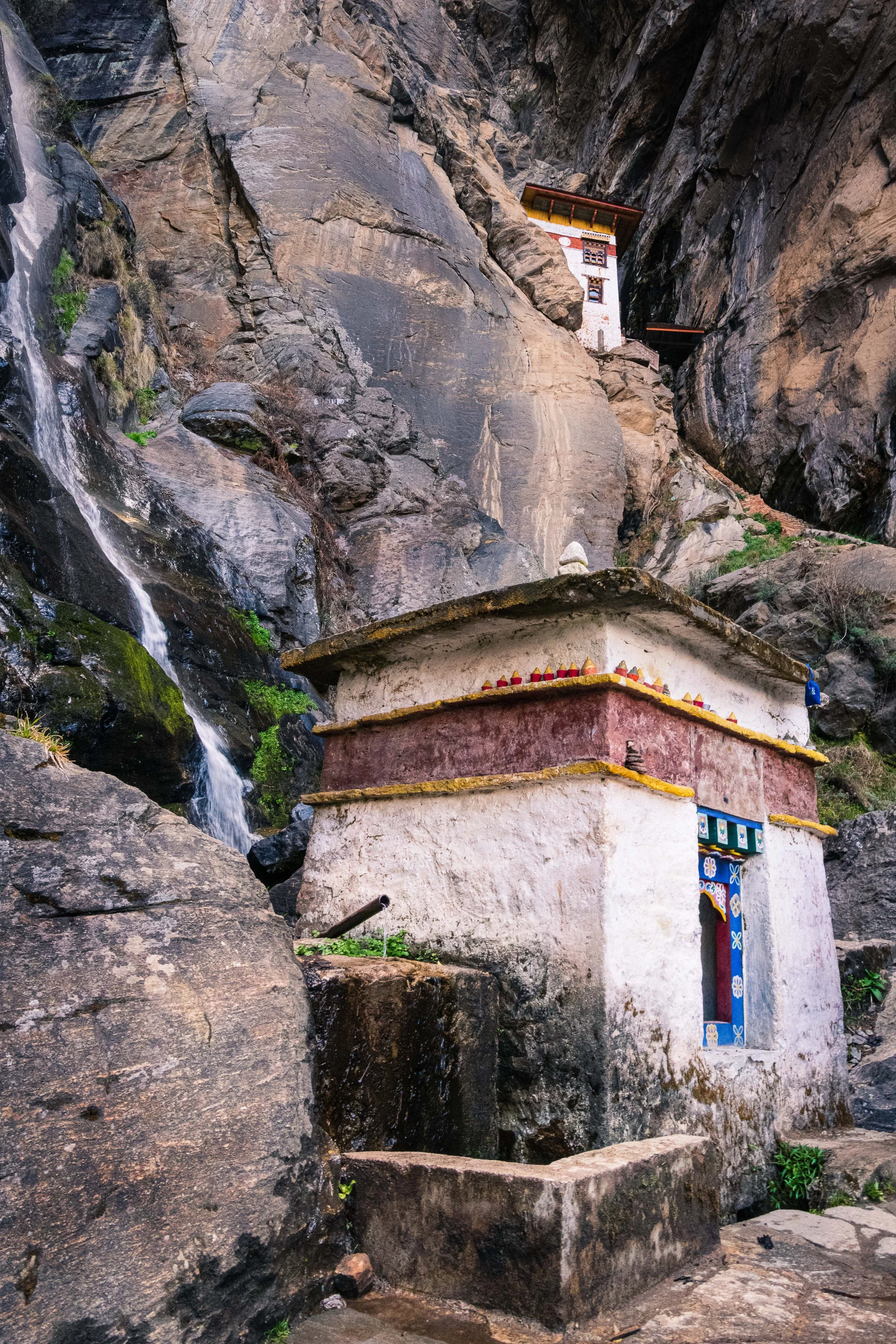 A small temple or shrine built against a rocky cliff with a waterfall nearby, featuring painted decorations and a small spout of water.