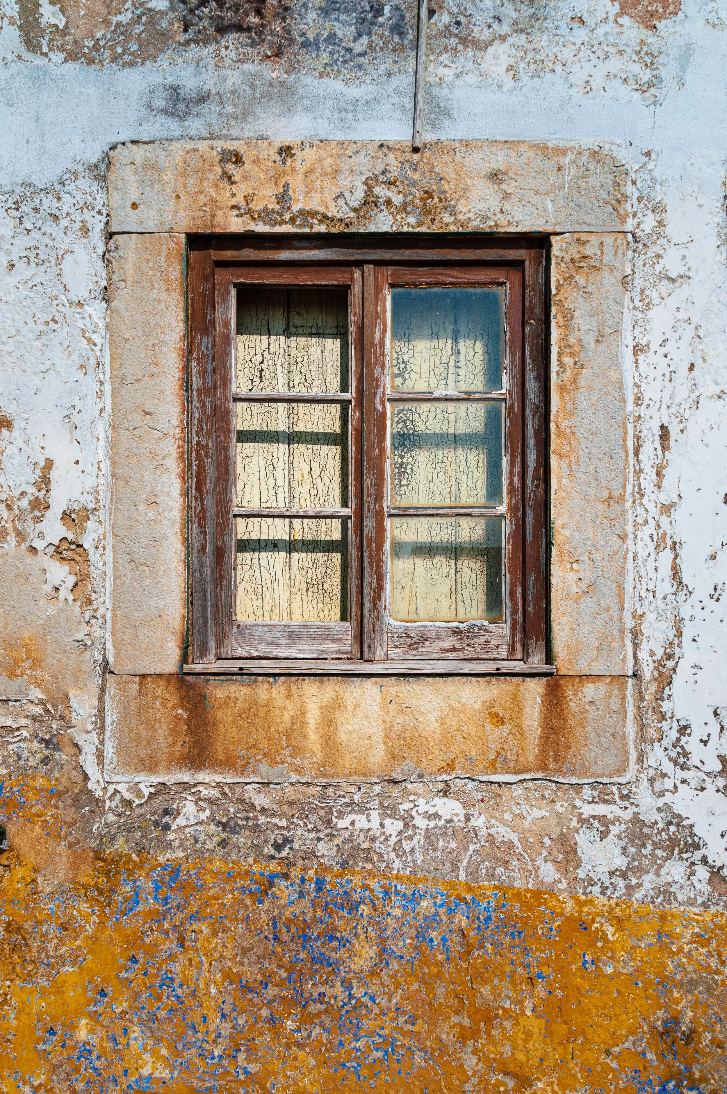 Old weathered wooden window with cracked glass set in a distressed stucco wall with peeling white and orange paint.