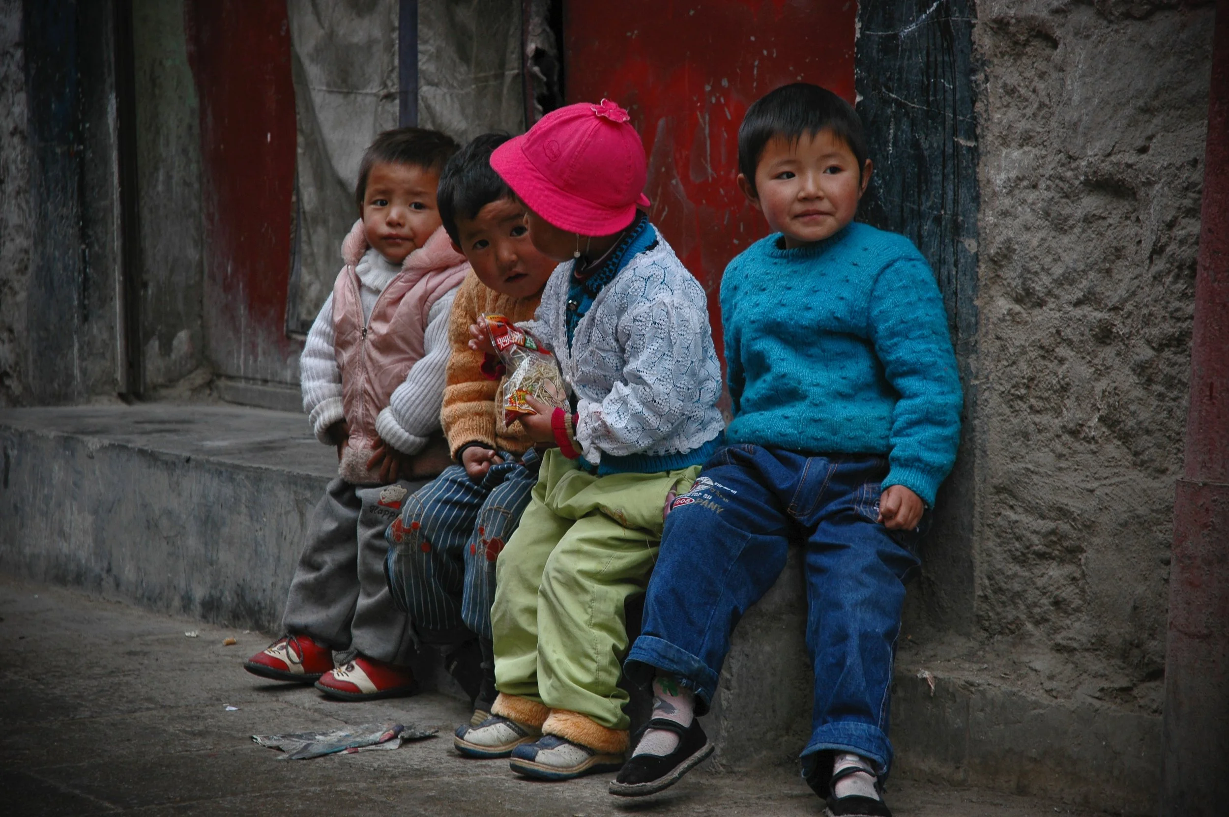 Five young children sitting on a ledge against a weathered wall, dressed in warm clothing, some holding snacks, looking in different directions.