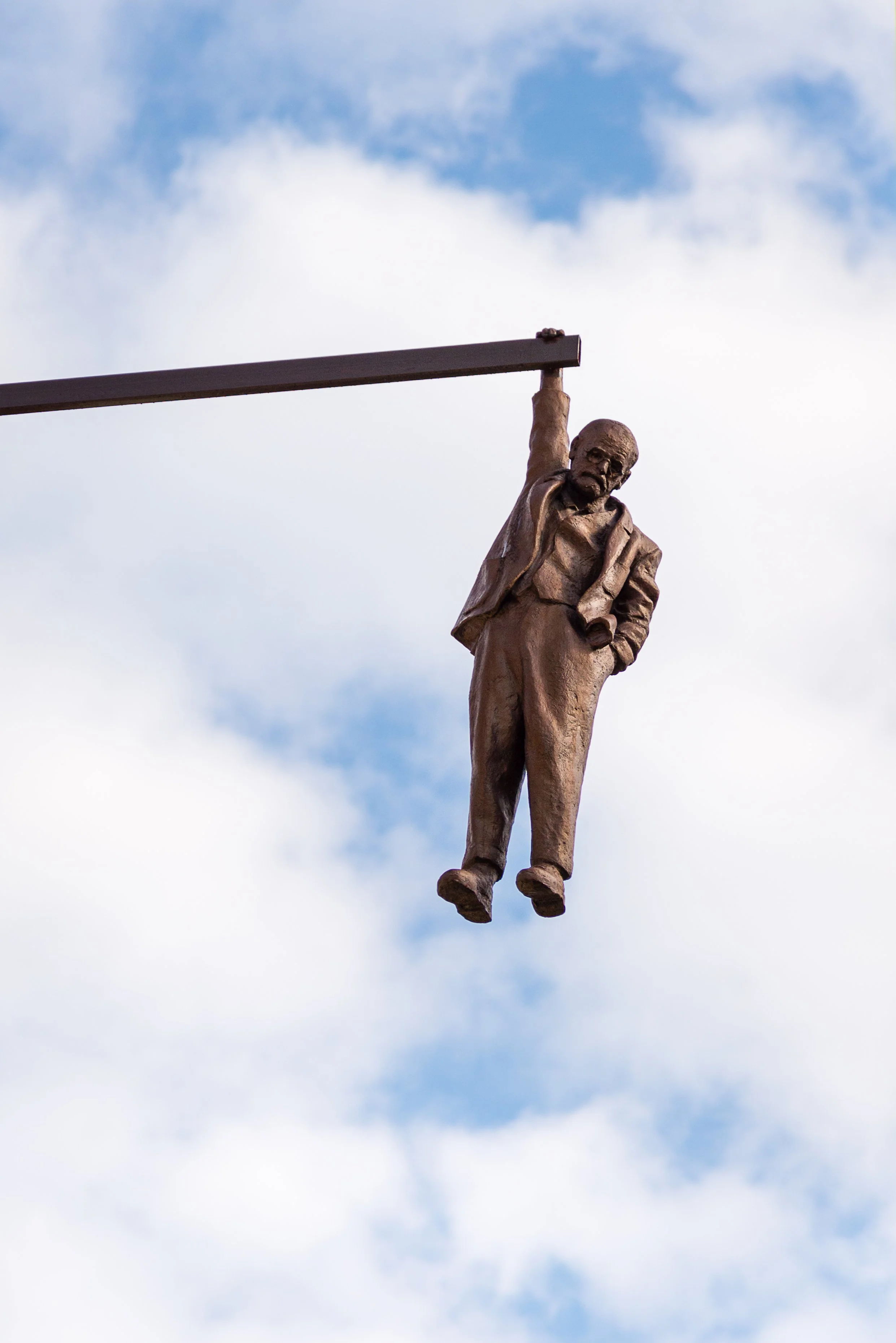 Statue of a man in a suit hanging from a pole against a cloudy sky.