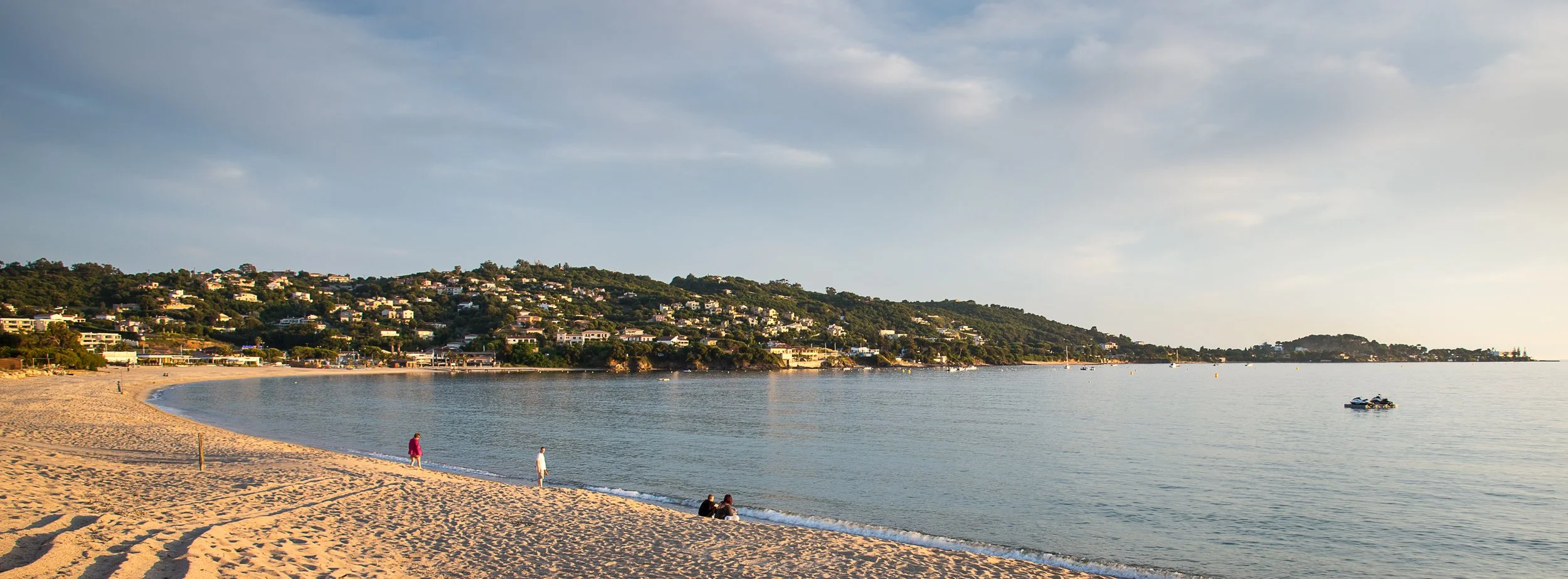 A beach scene with a sandy shoreline, calm water, a hillside with residential houses, and a few people walking and sitting near the water, with boats in the distance.