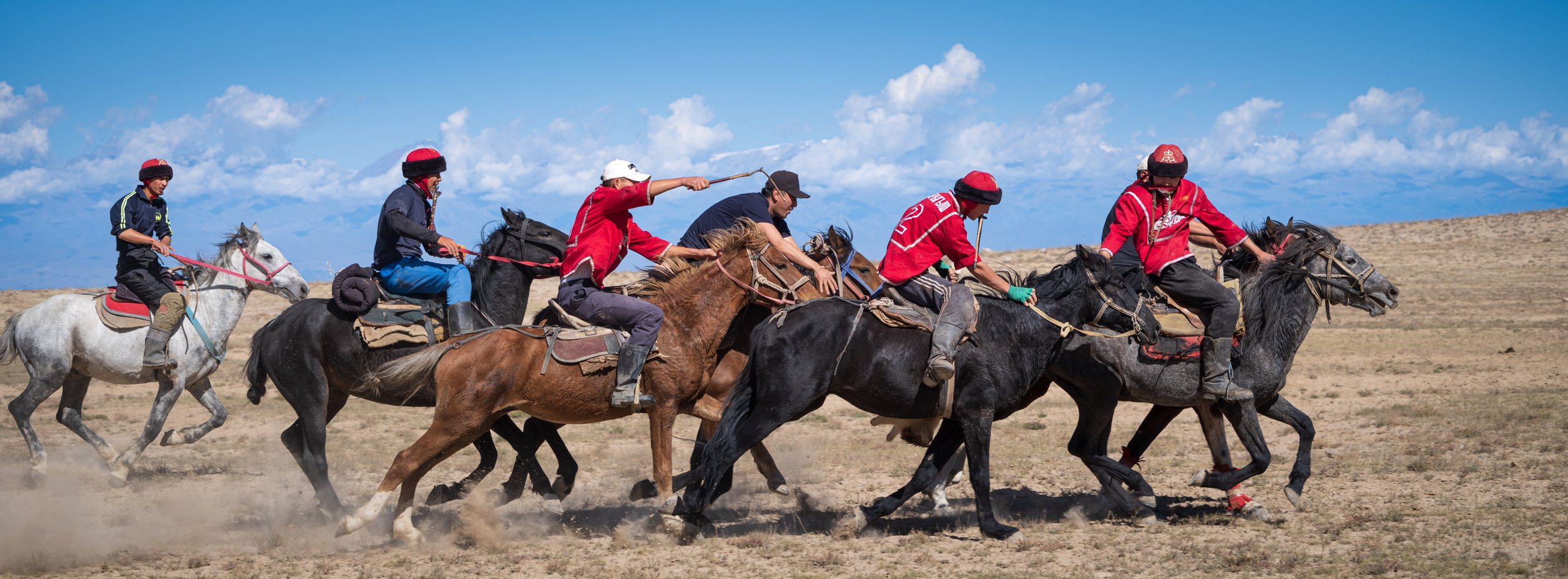 A group of people riding galloping horses in an open field under a blue sky with scattered clouds