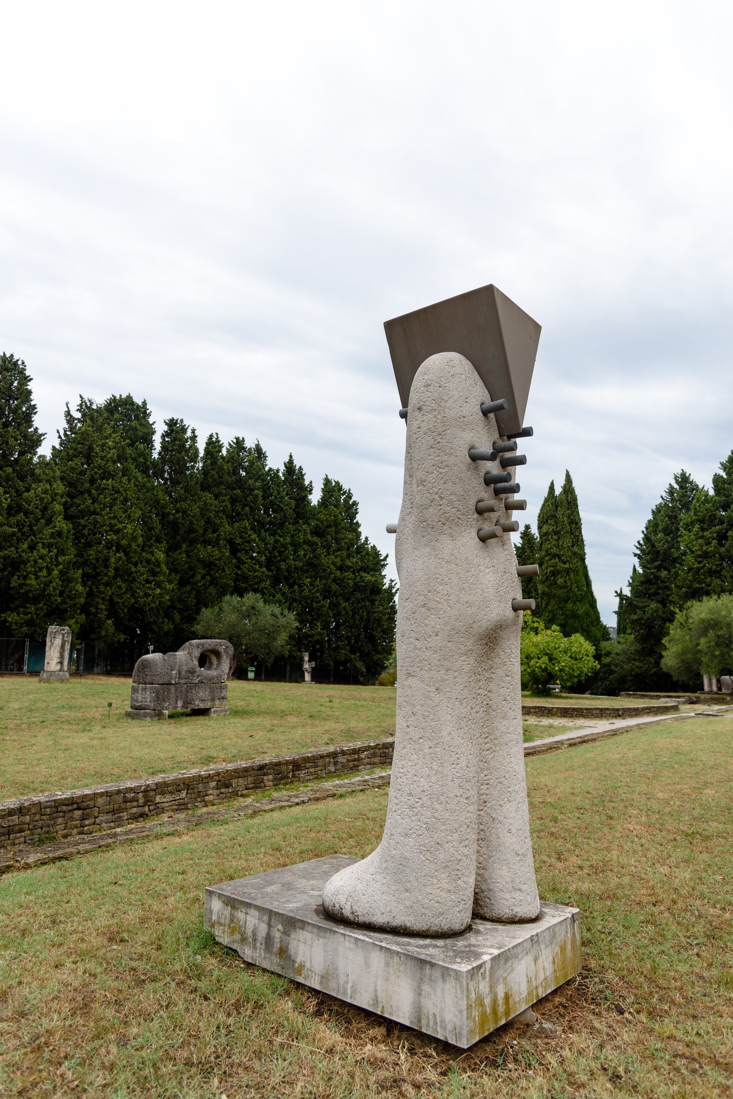 Abstract modern outdoor sculpture resembling a pair of large beige legs with irregular texture, with a dark rectangular shape on top and metal rods protruding from the sides, situated in a grassy park with trees in the background.