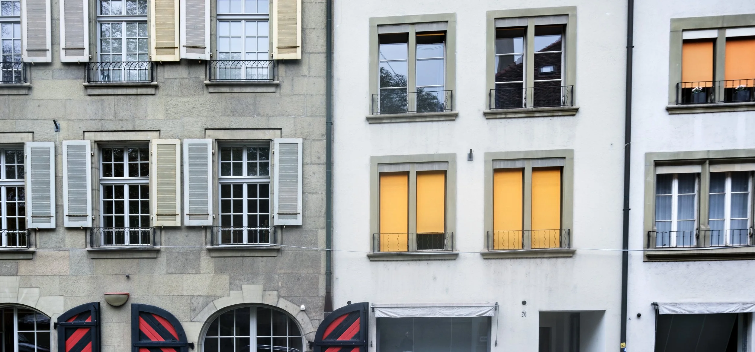 Close-up of three-story residential buildings with windows, some with shutters, some with blinds, and small balconies, in an urban setting.