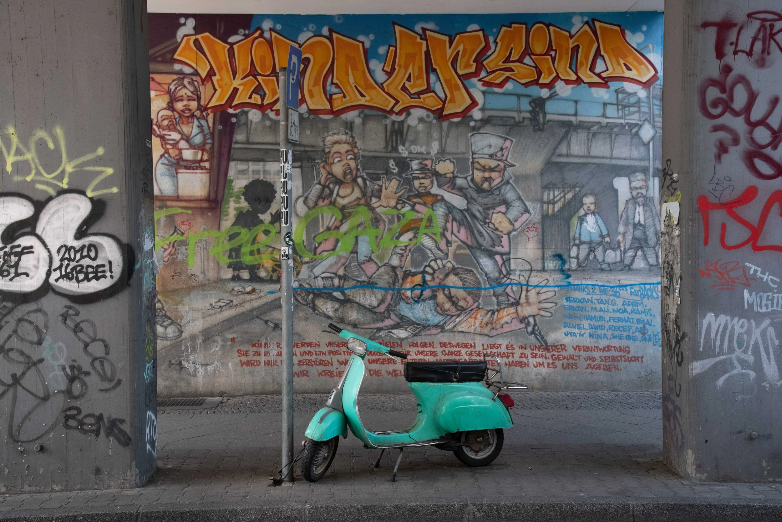 Graffiti mural depicting a chaotic scene with people and dogs, and a turquoise scooter parked in front under a covered structure.