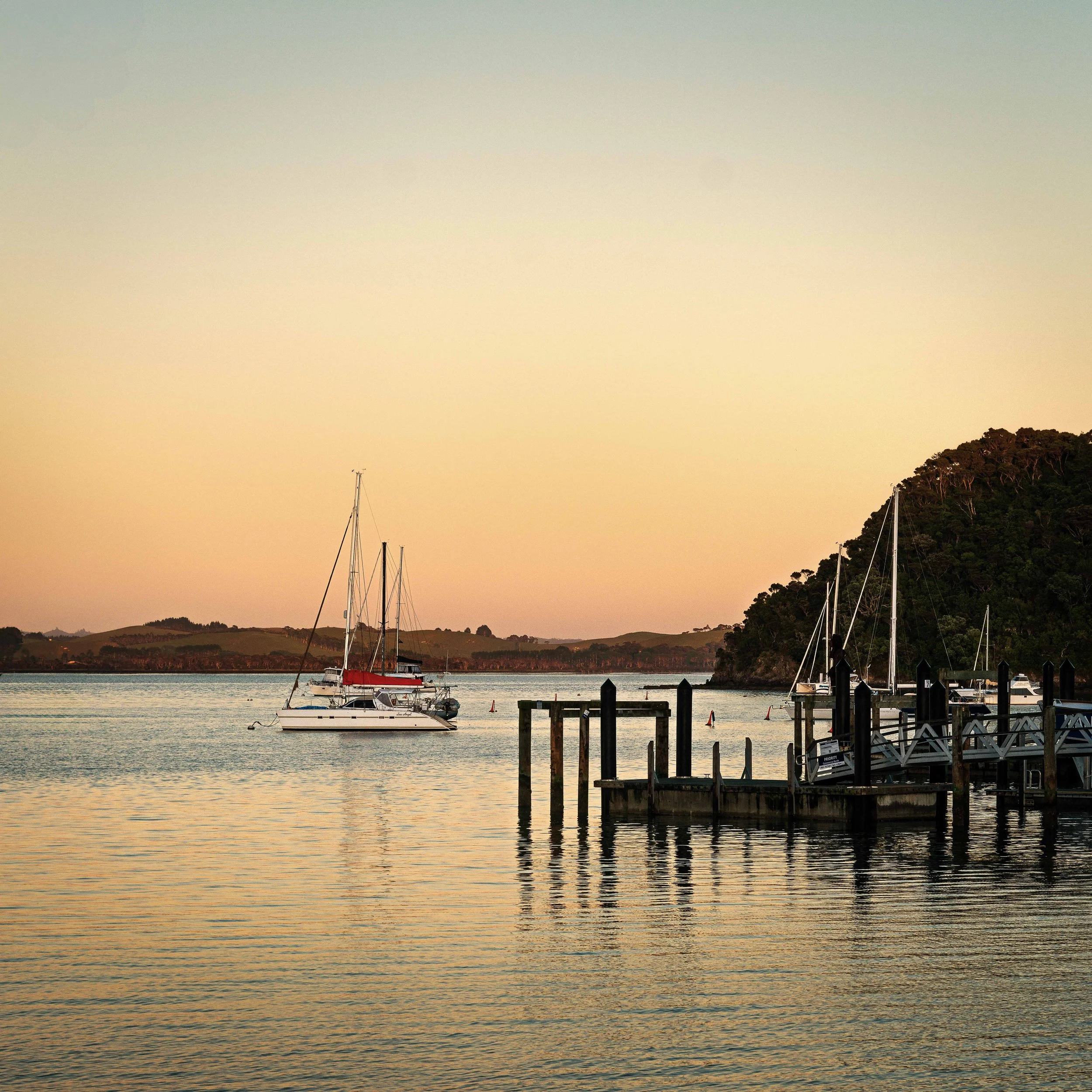 Calm water with three sailboats docked near a pier at sunset, with a forested hill in the background.
