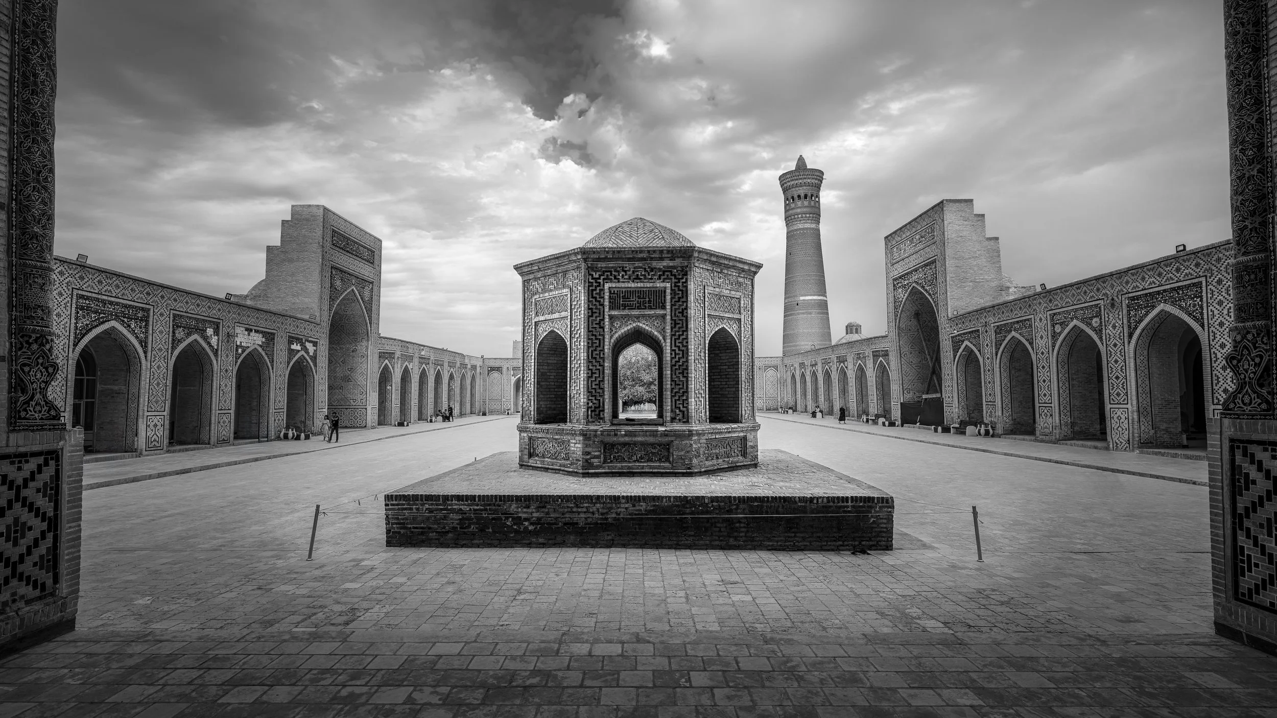 Black and white photo of an ancient architectural courtyard with intricate tile designs, arches, and a tall minaret in the background, with a central pavilion structure.