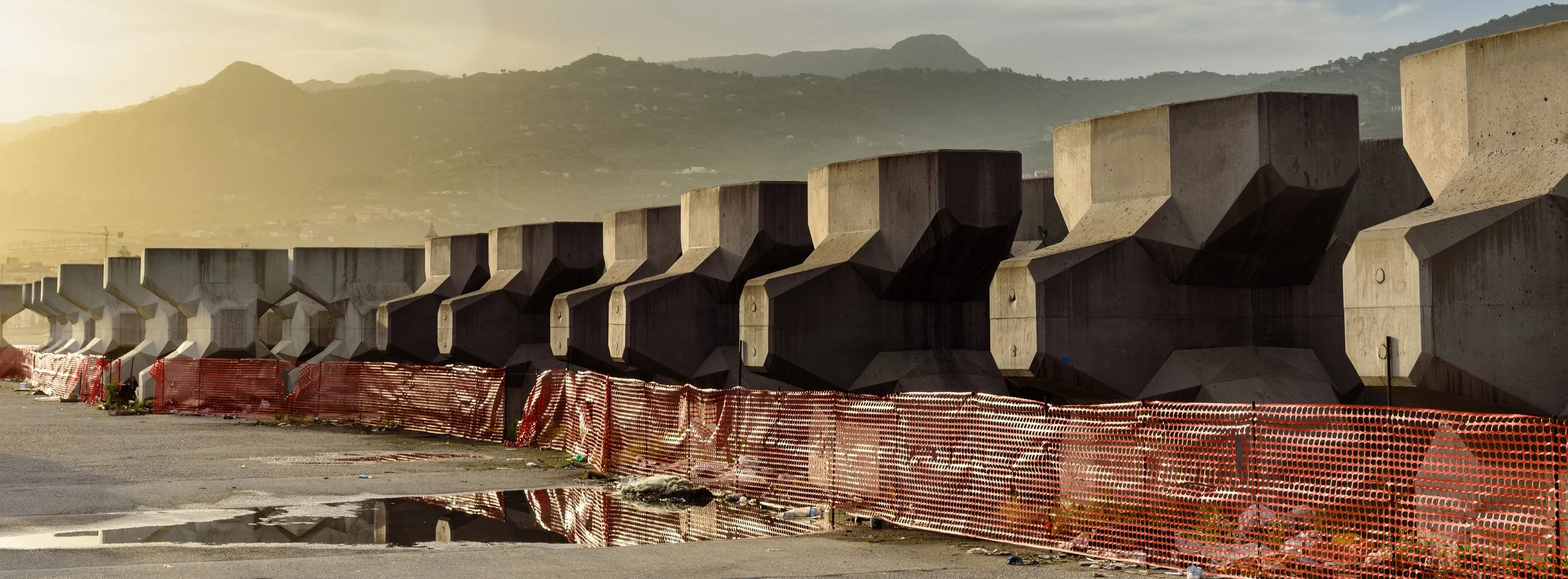 Concrete barriers along a construction site with orange safety fencing, puddles on the ground, under a cloudy sky with mountains in the background.
