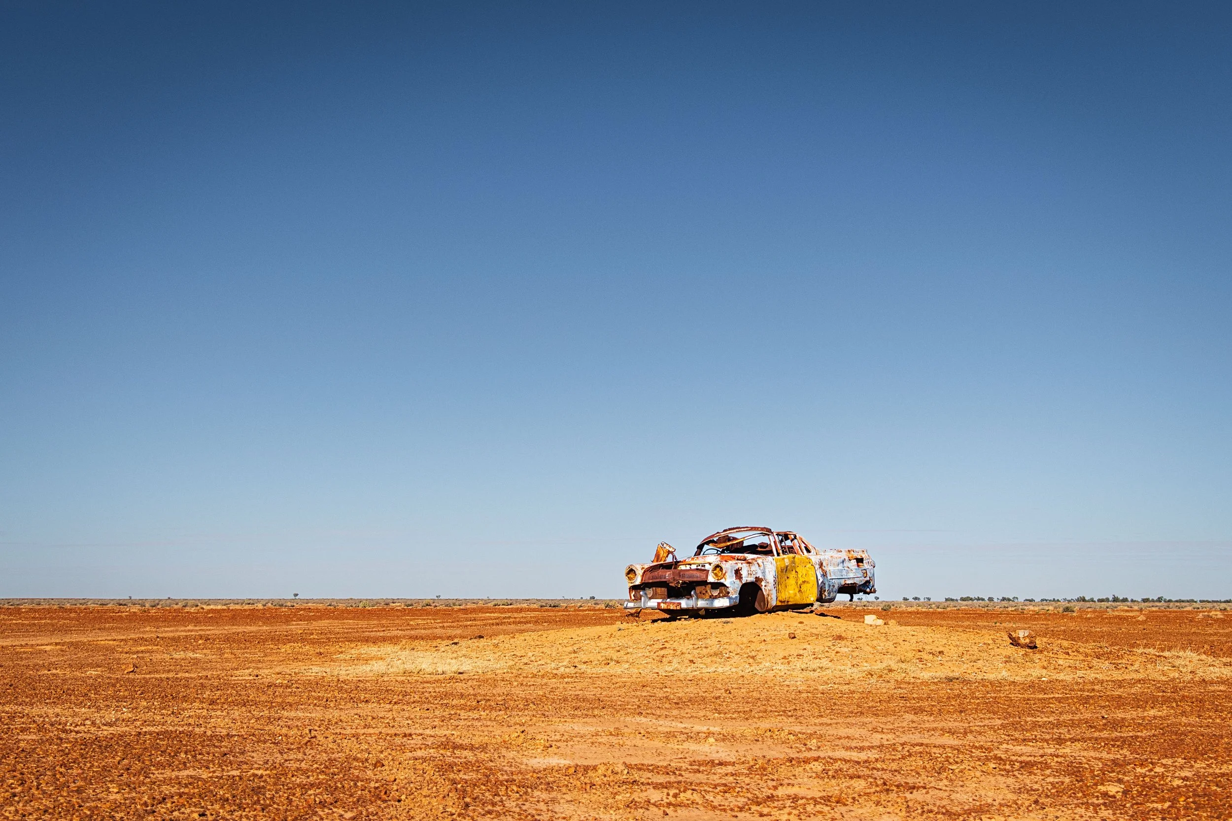 Rusty, abandoned car in a barren desert landscape under a clear blue sky.